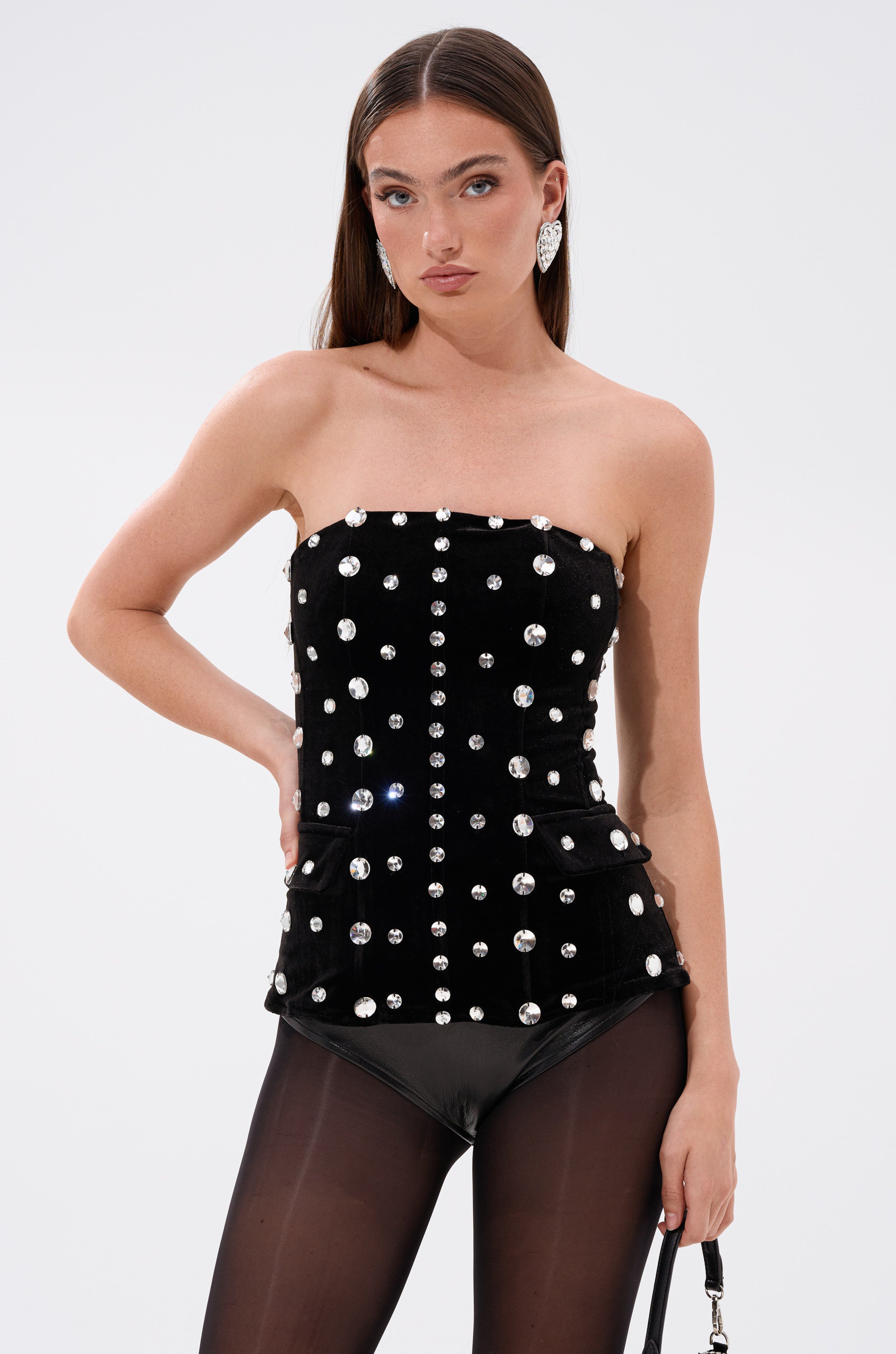 A woman with long brown hair and statement earrings poses confidently in the CLASSY GIRL VELVET CORSET WITH RHINESTONE, featuring rows of silver studs and rhinestones, paired with black tights. 0