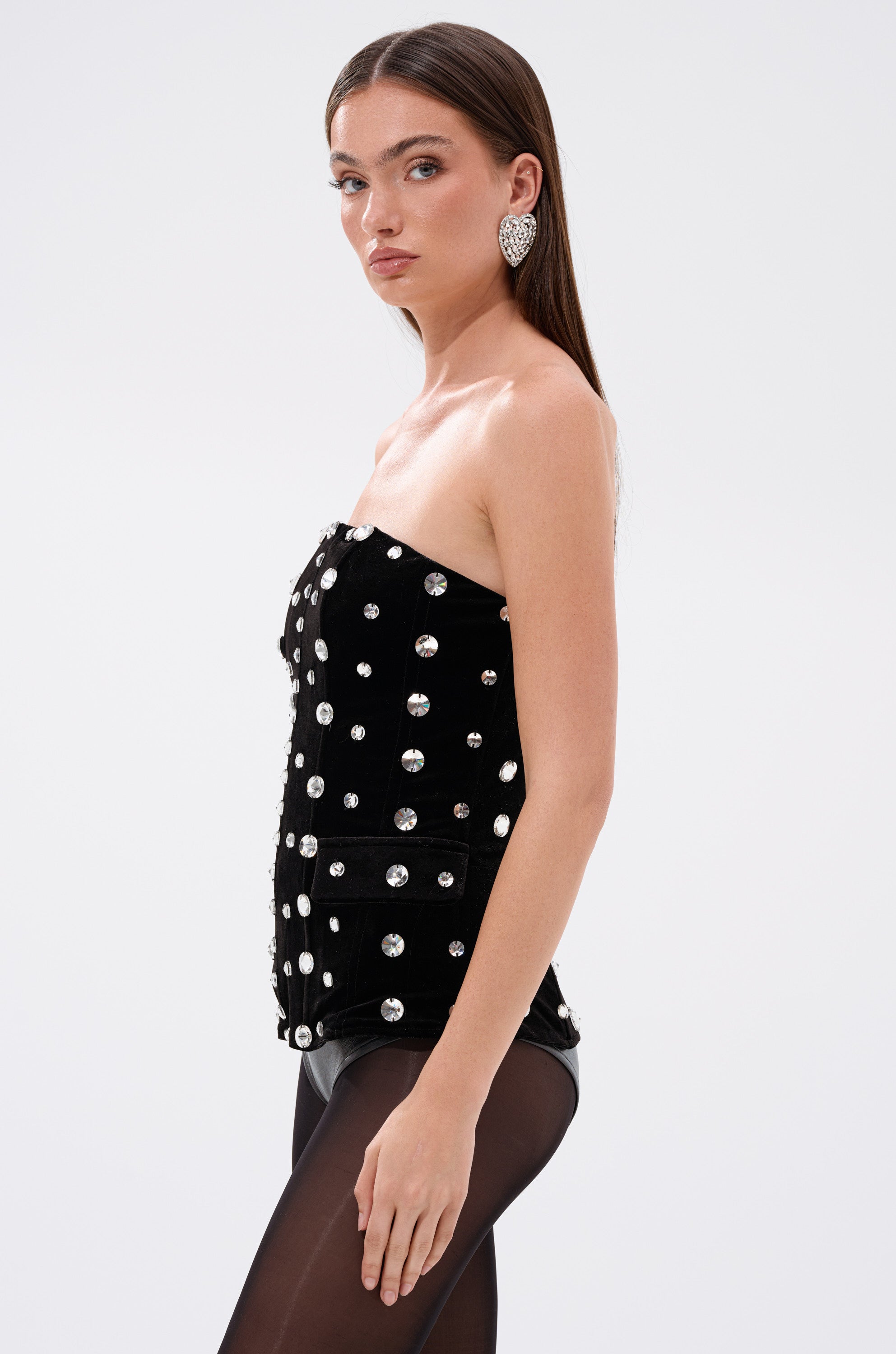A woman with straight brown hair models the CLASSY GIRL VELVET CORSET WITH RHINESTONE, paired with sheer black tights and silver statement earrings, posing against a plain white background. 3
