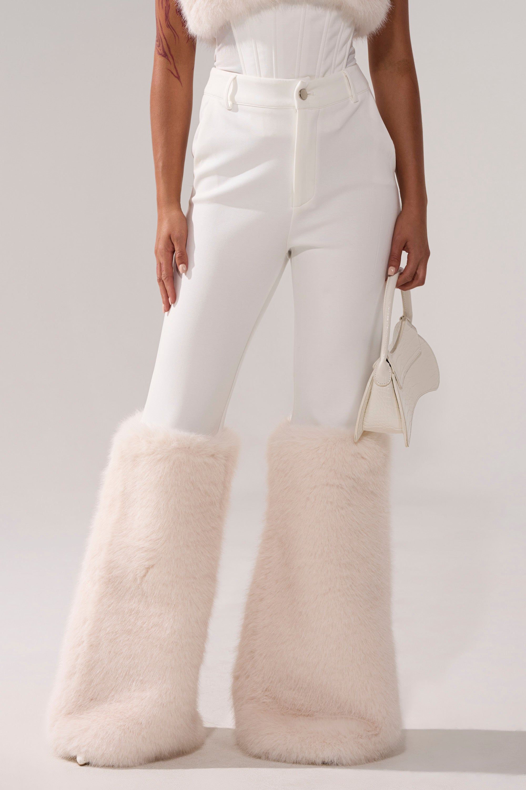 Someone stands against a light background wearing high-waisted SNOW BUNNY TROUSER WITH FUR CUFF, featuring large, fluffy, pale pink faux fur leg cuffs and holding a small white handbag.