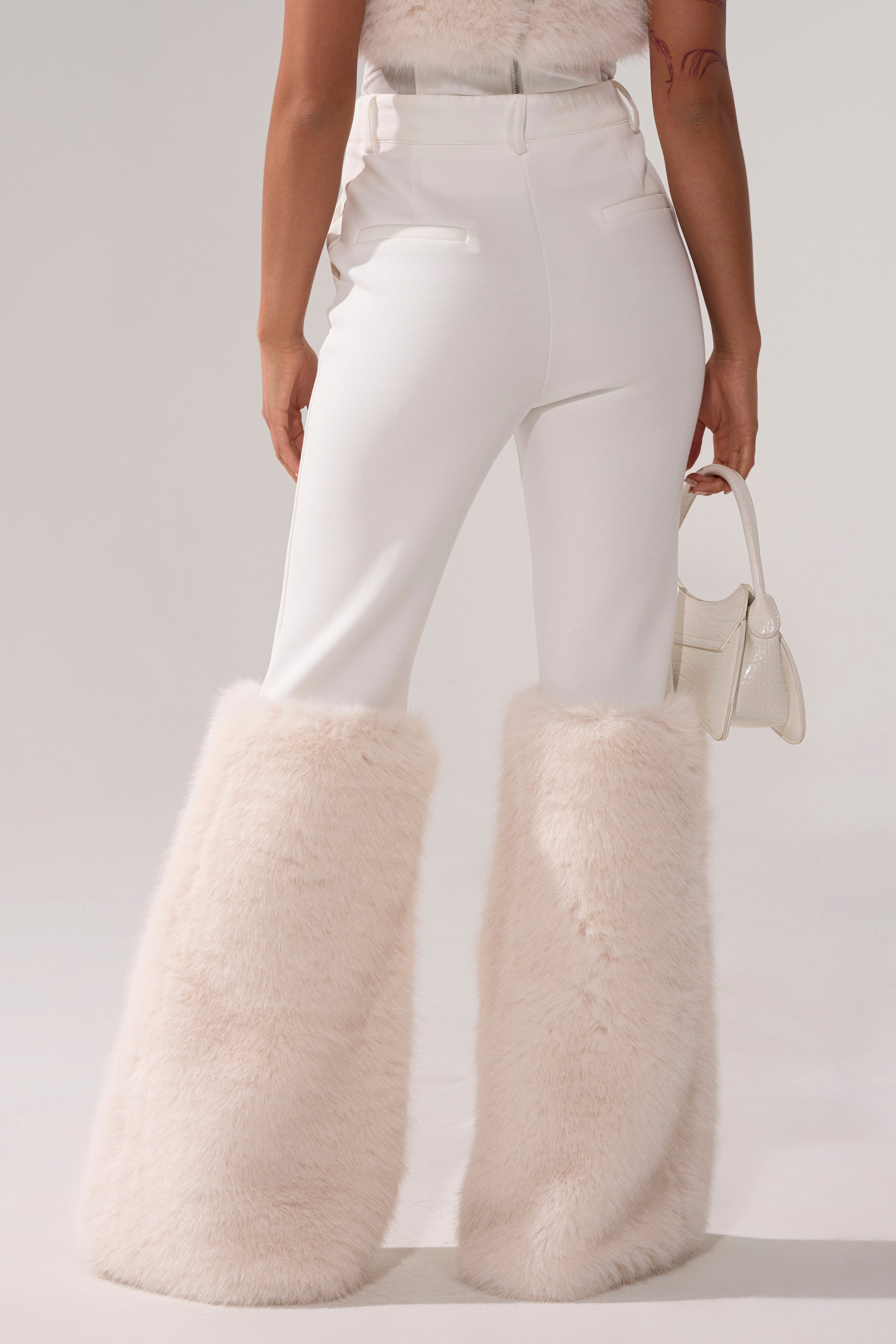 A person wearing the SNOW BUNNY TROUSER WITH FUR CUFF—featuring large, fluffy, pale pink cuffs—holds a small white handbag and is photographed from behind against a plain background.