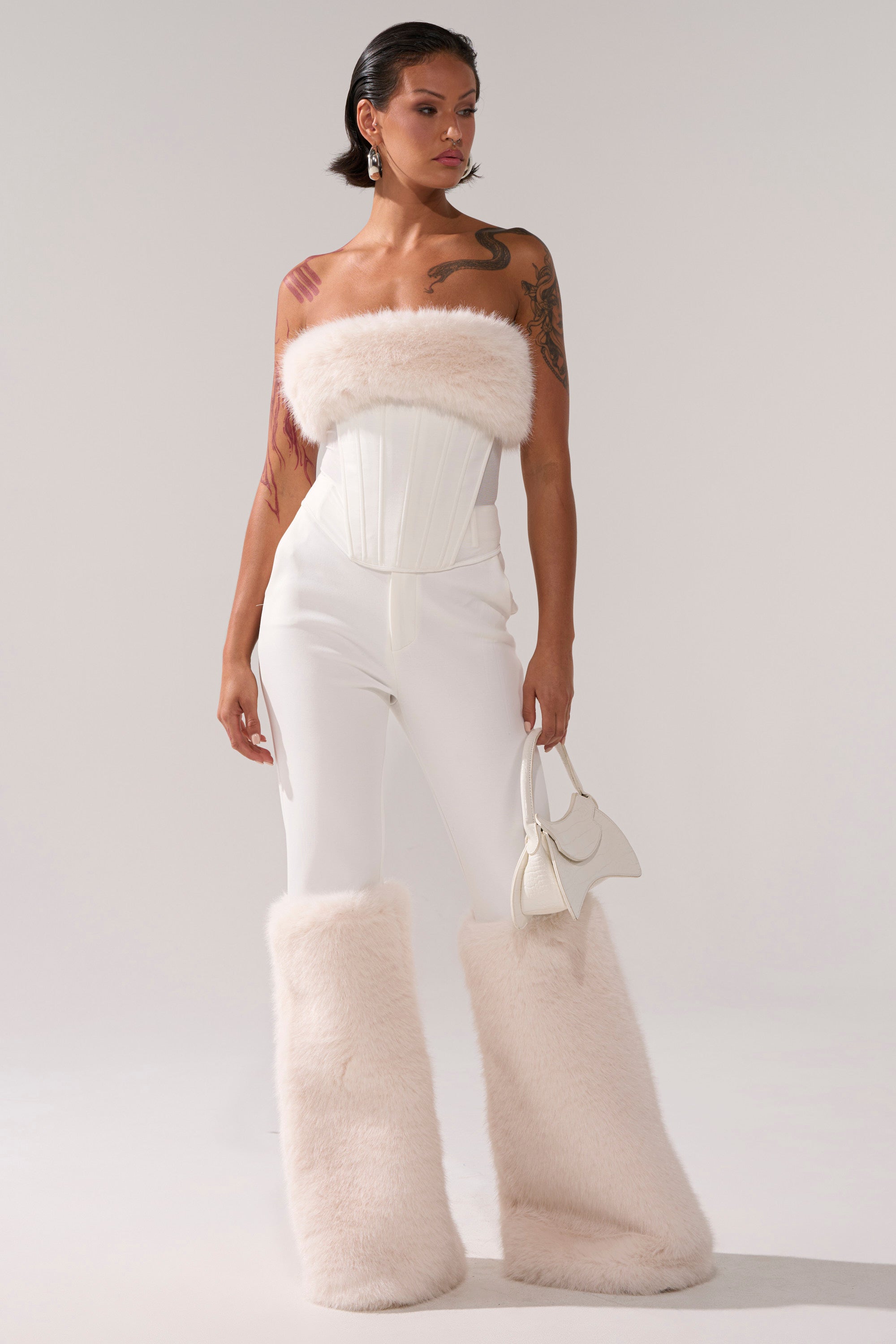 A woman in a white strapless top, SNOW BUNNY TROUSER WITH FUR CUFF featuring large faux fur leg warmers, and holding a small white handbag poses against a plain light background.