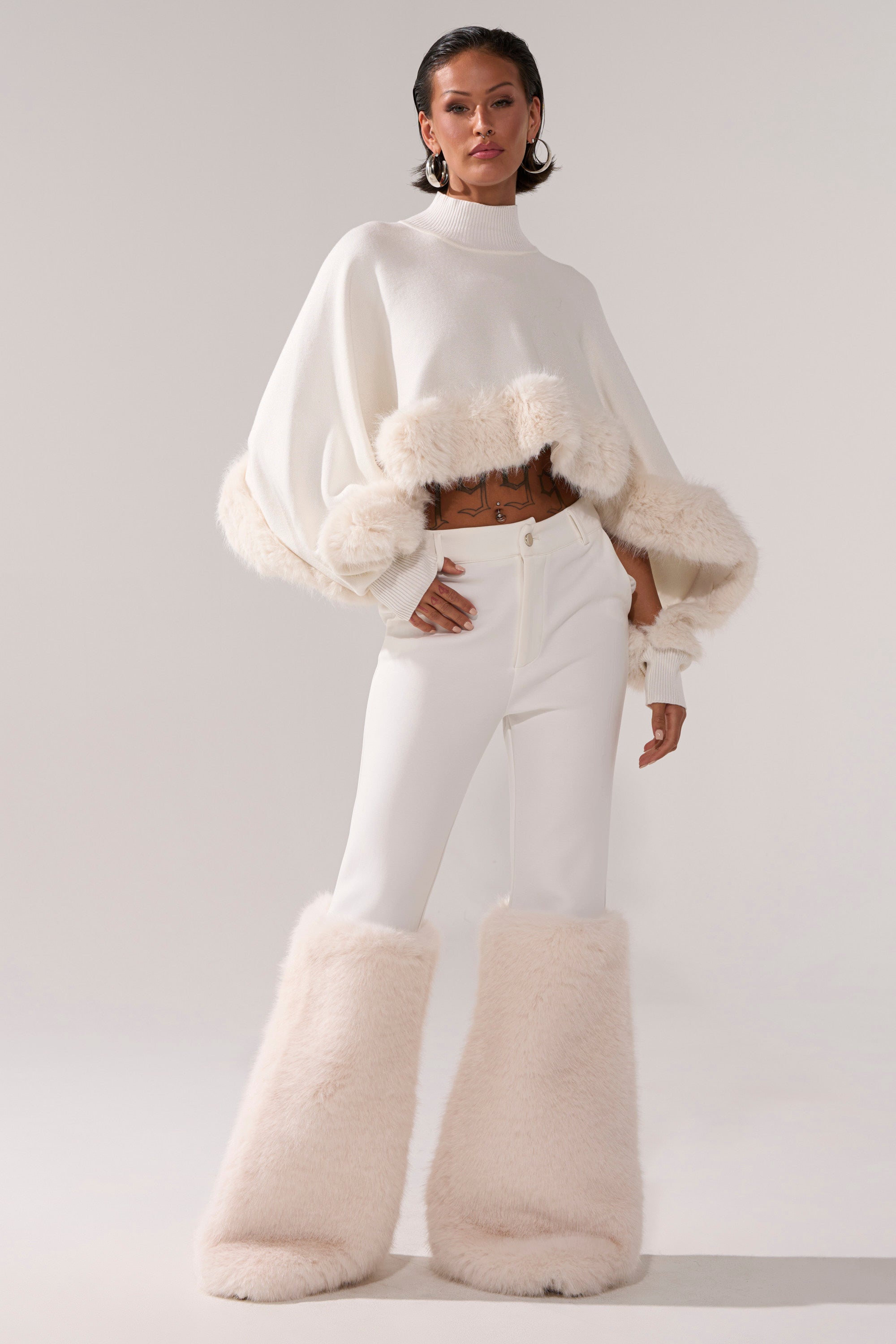 A person stands confidently in a white sweater, SNOW BUNNY TROUSER WITH FUR CUFF, and oversized fluffy white boots, creating a chic and bold fashion-forward look against a plain background.