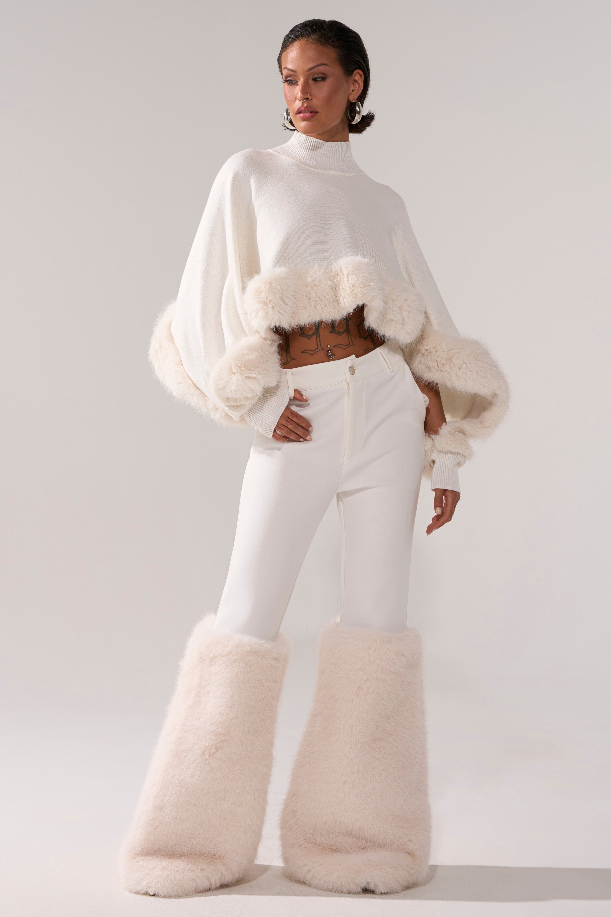 A person poses confidently in an all-white outfit featuring a cropped sweater with fluffy trim, SNOW BUNNY TROUSER WITH FUR CUFF, and tall furry boots against a plain light-colored background.