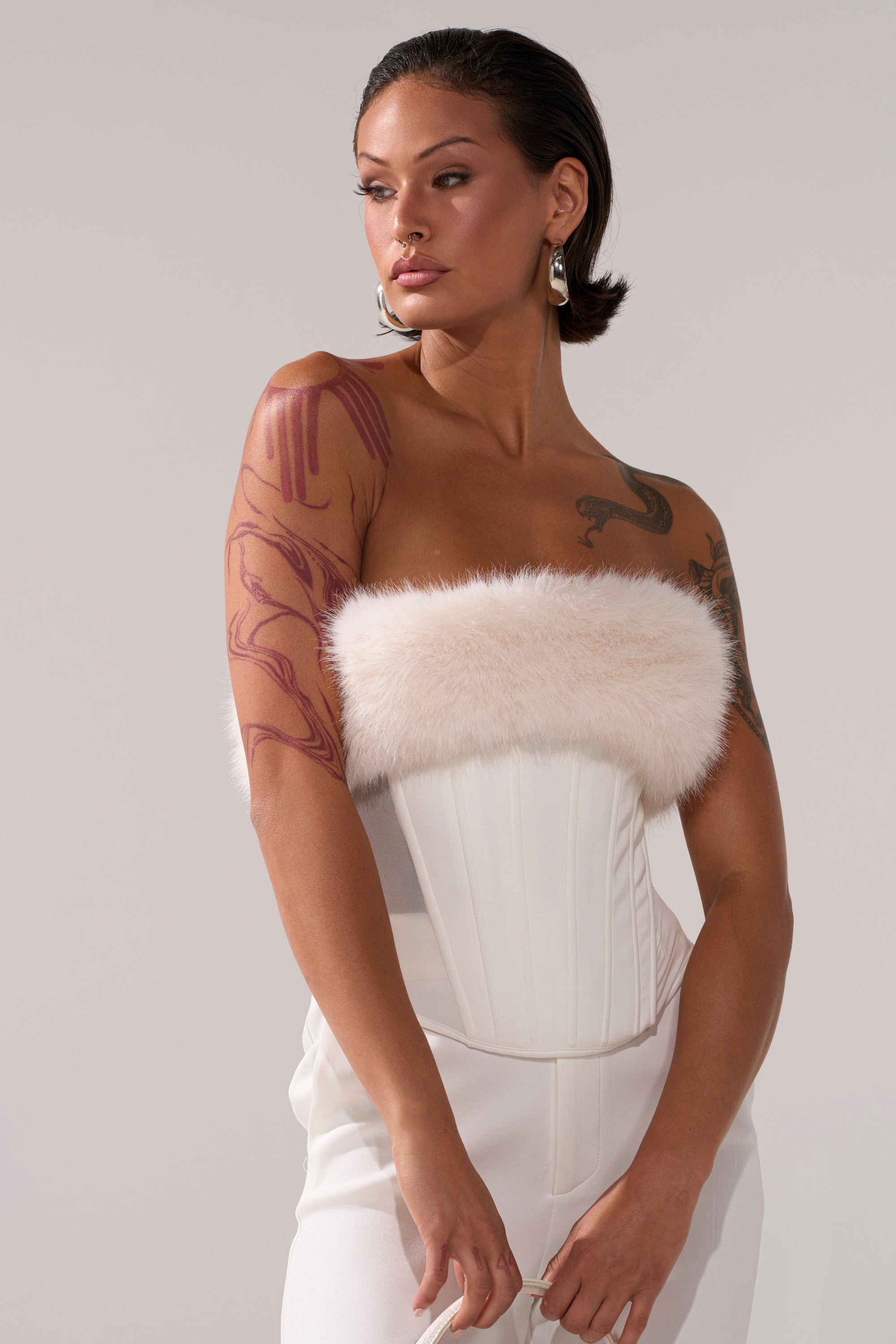 A person with short dark hair, bold earrings, and arm tattoos models the SNOW BUNNY CORSET WITH FUR and white pants, gazing confidently to the side against a plain background.