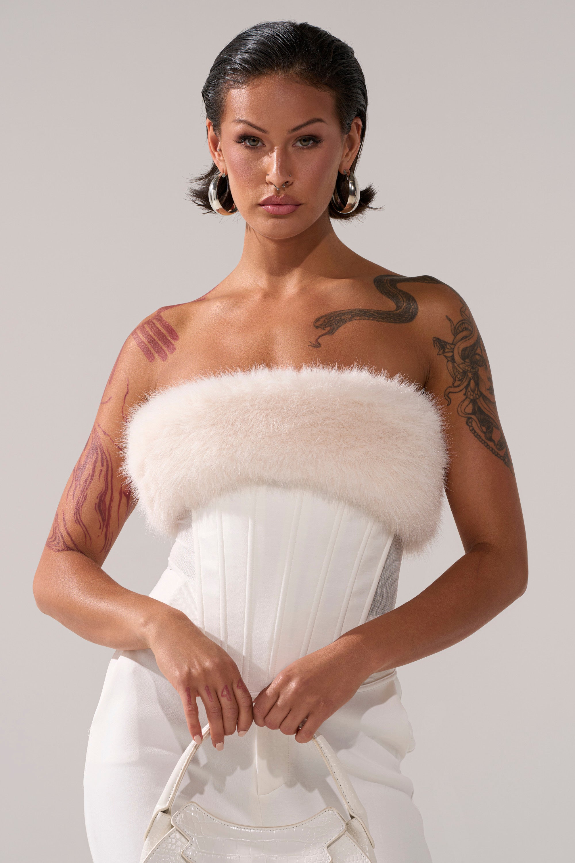 A woman with tattoos and short dark hair wears the SNOW BUNNY CORSET WITH FUR and white pants, accessorized with a small white handbag. She poses confidently against a plain background.