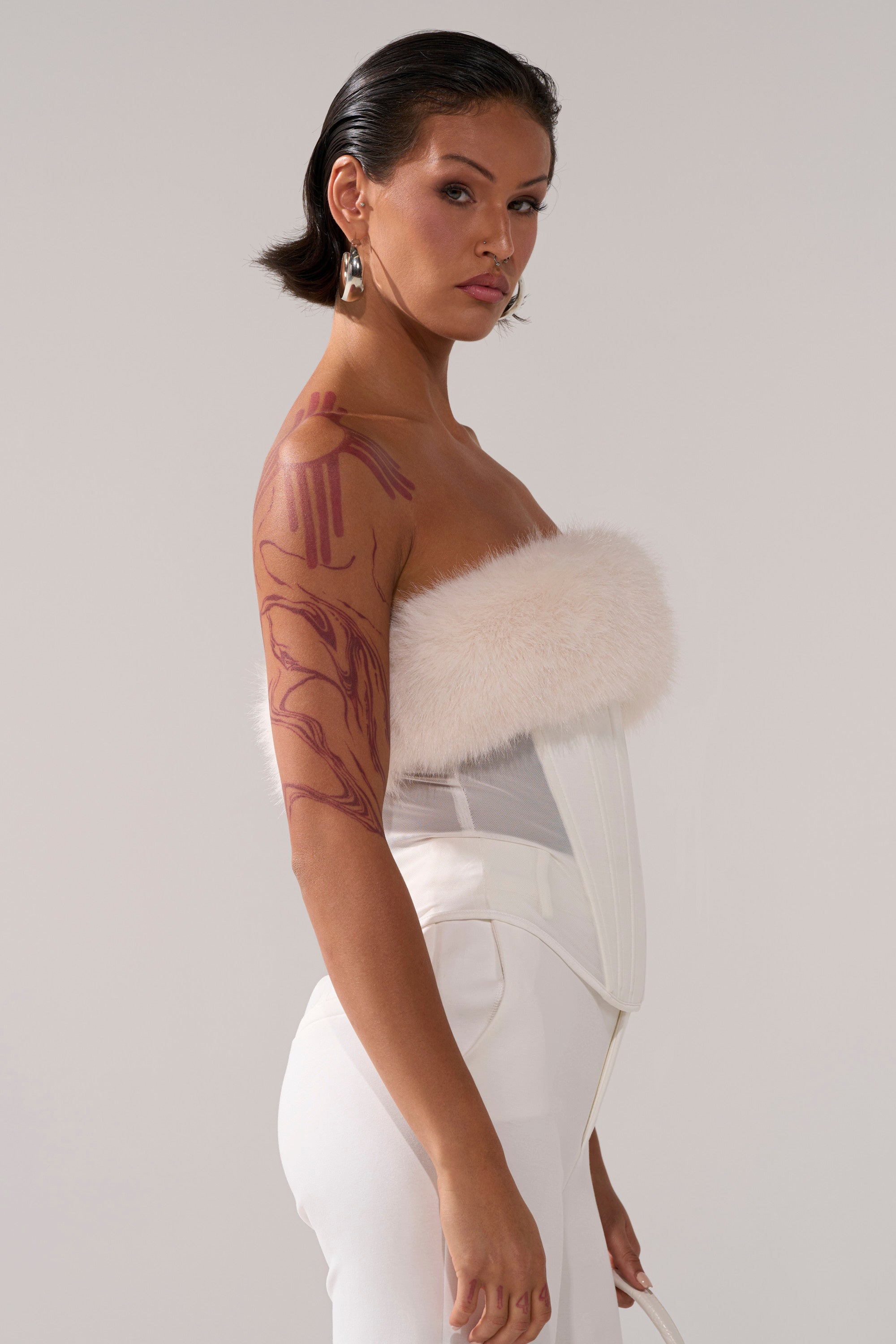 A woman stands sideways in the SNOW BUNNY CORSET WITH FUR, featuring an off-shoulder fluffy trim and white pants. She has slicked-back dark hair, silver earrings, and visible red body art on her arm against a plain light background.
