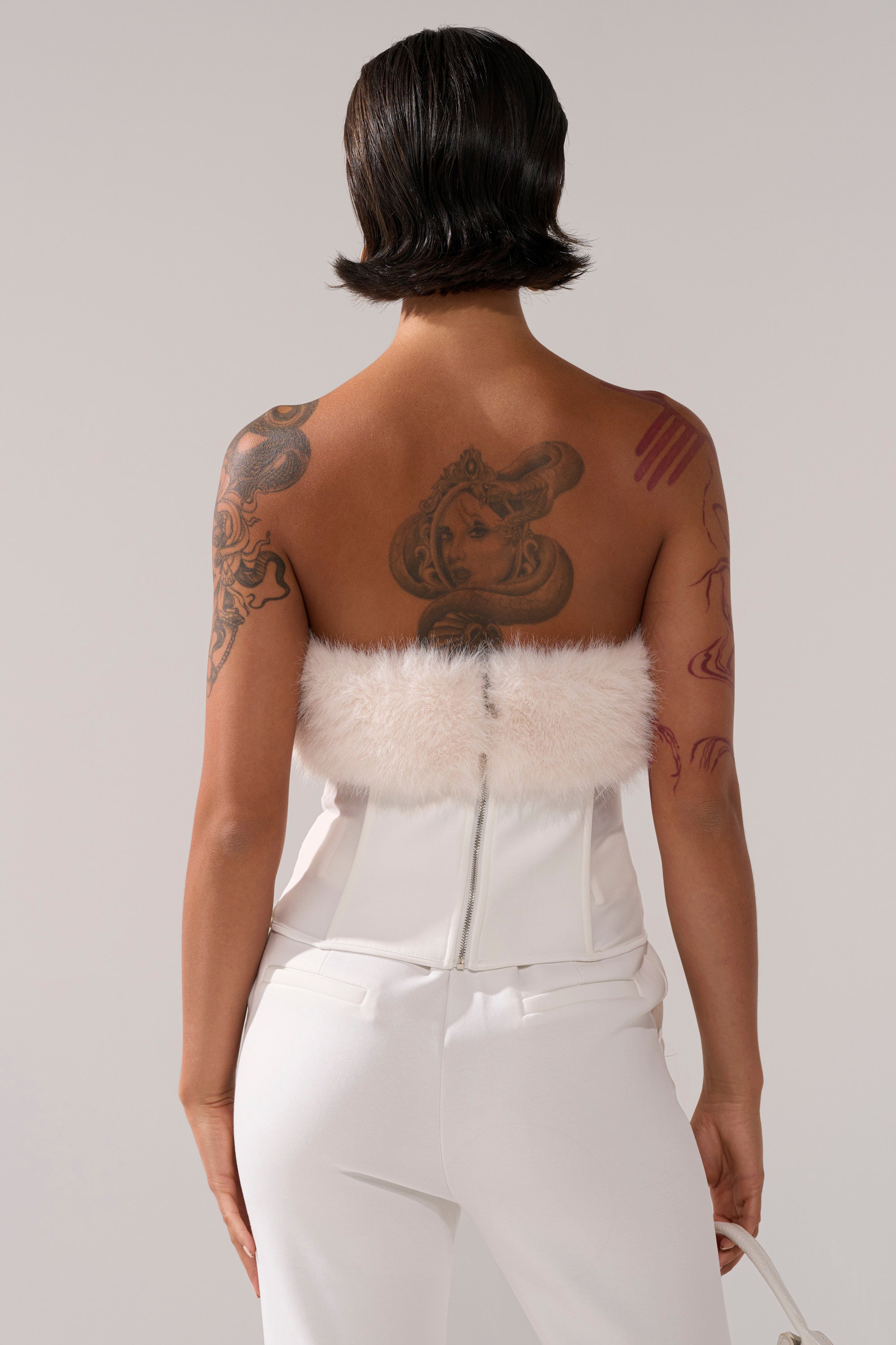 A person with short dark hair and visible tattoos is seen from behind in a strapless SNOW BUNNY CORSET WITH FUR and white pants, set against a plain background.