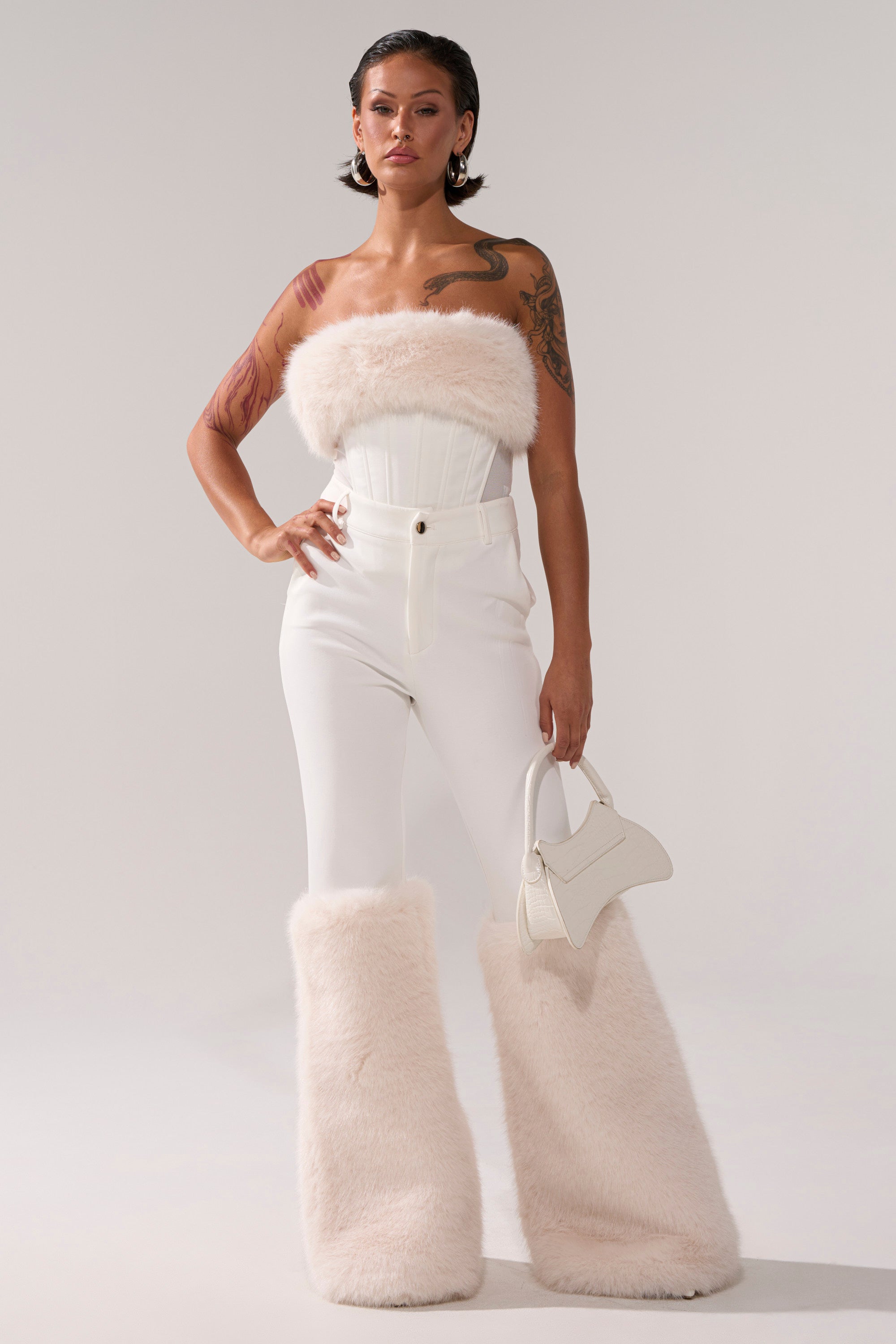 A woman stands confidently wearing the SNOW BUNNY CORSET WITH FUR, paired with high-waisted pants, matching furry leg warmers, and holding a small white handbag against a plain background.