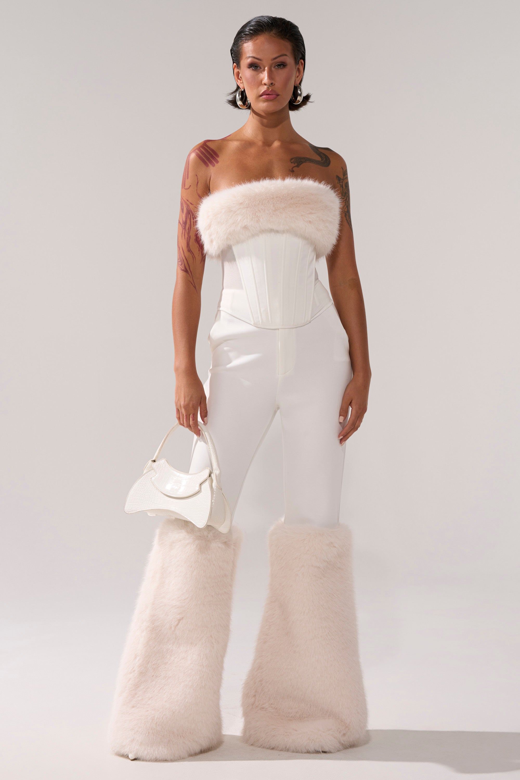 A woman with tattoos and short, slicked-back hair models the SNOW BUNNY CORSET WITH FUR, paired with fitted white pants featuring fur at the lower legs and a white handbag, standing against a plain background.