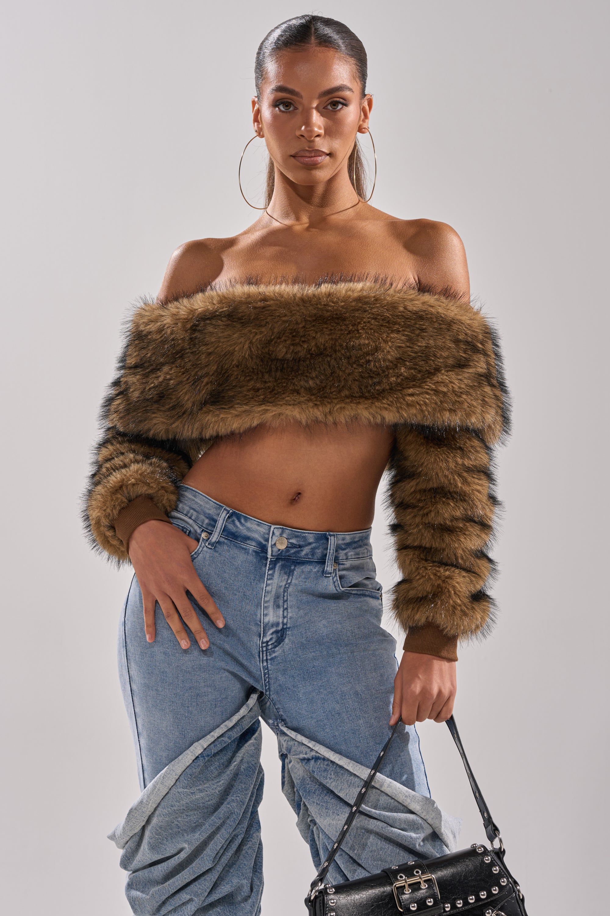 A woman confidently wears the TEDDY BEAR CROPPED LONG SLEEVE TOP off the shoulder, pairing it with high-waisted light blue jeans featuring twisted seams, a black studded handbag, and large hoop earrings.