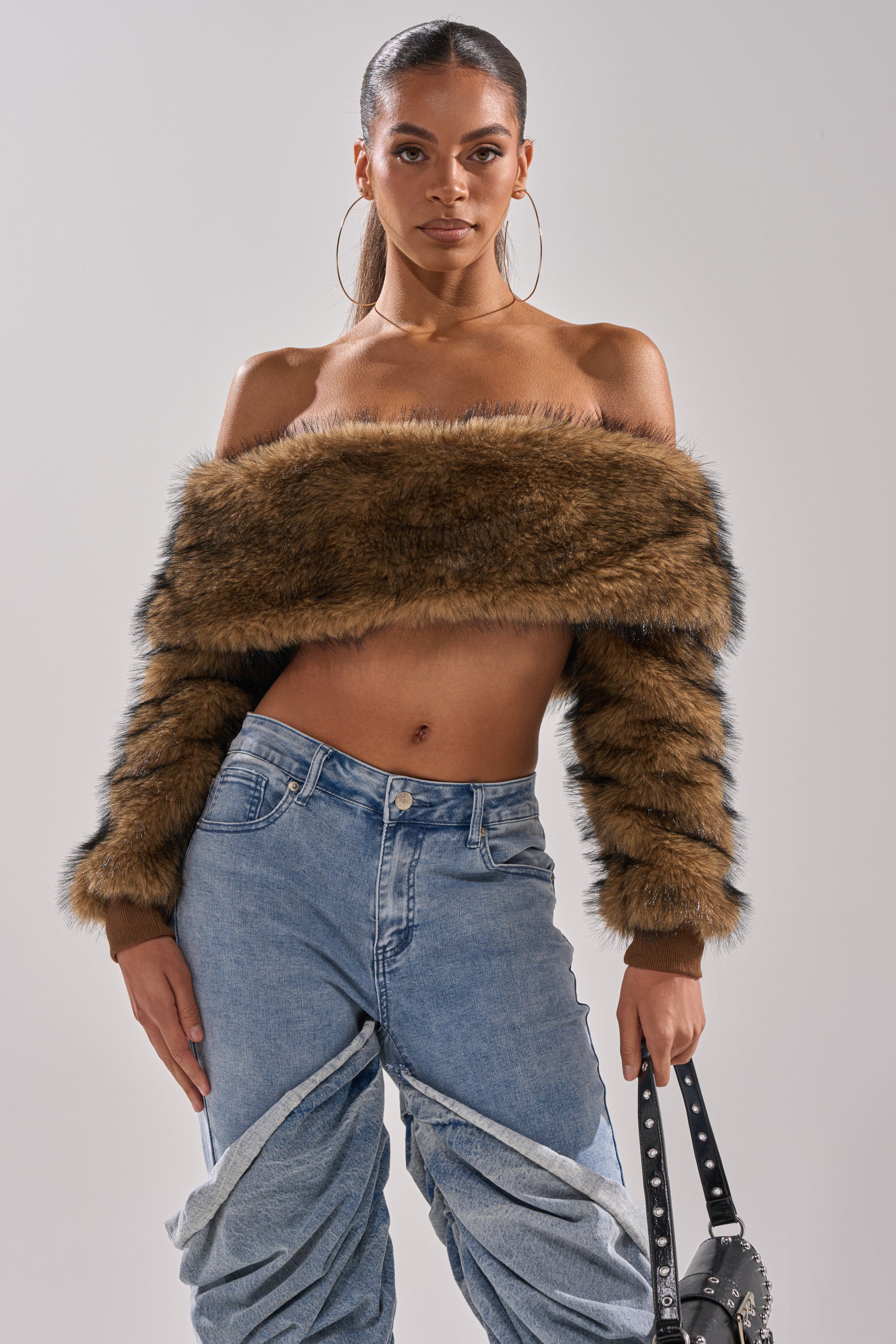 A woman wears the TEDDY BEAR CROPPED LONG SLEEVE TOP in brown faux fur, paired with high-waisted blue jeans, large hoop earrings, and holds a black studded purse while standing against a neutral background.