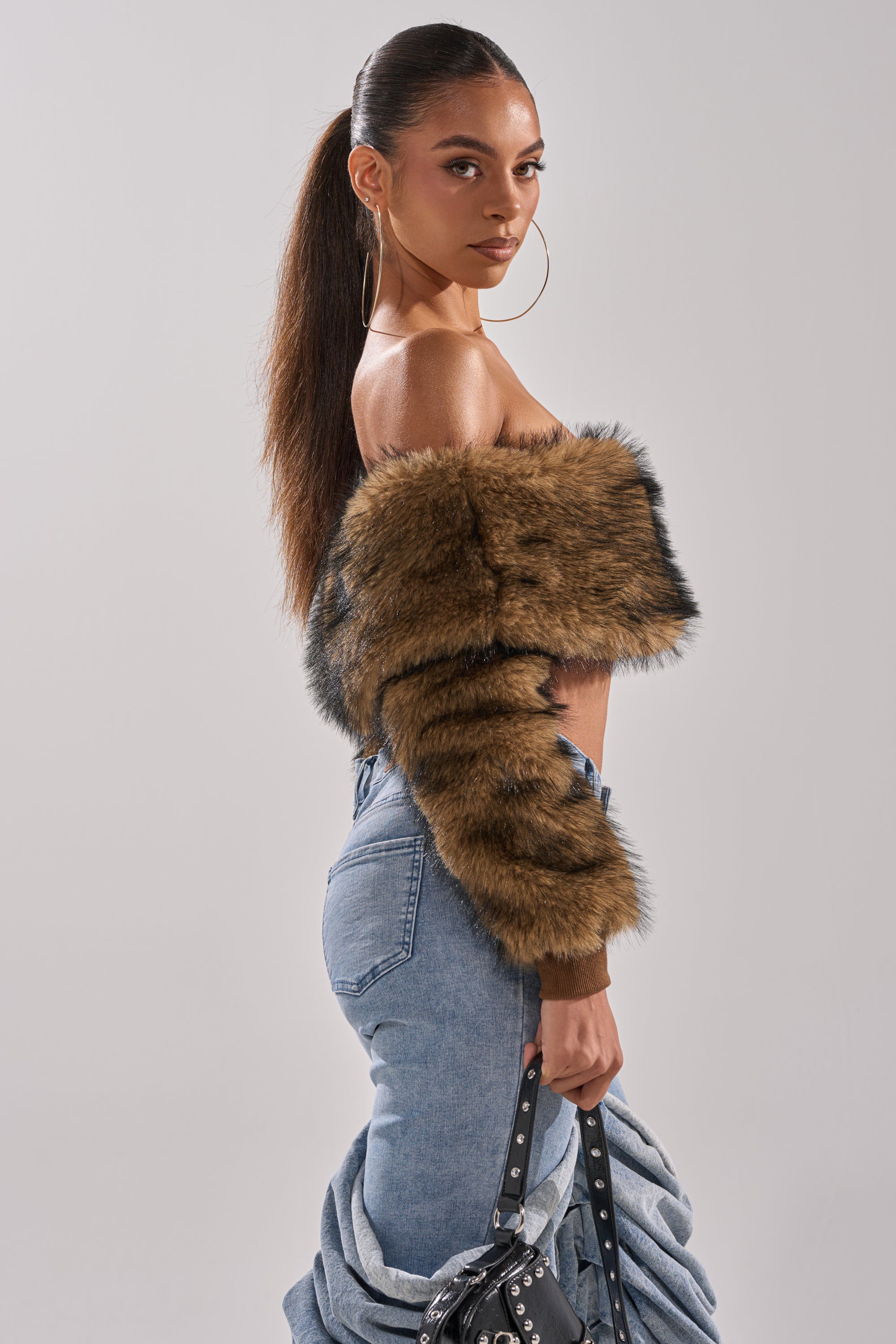 A woman with long dark hair in a high ponytail wears large hoop earrings, high-waisted jeans, a studded black handbag, and a TEDDY BEAR CROPPED LONG SLEEVE TOP, while carrying a denim jacket.