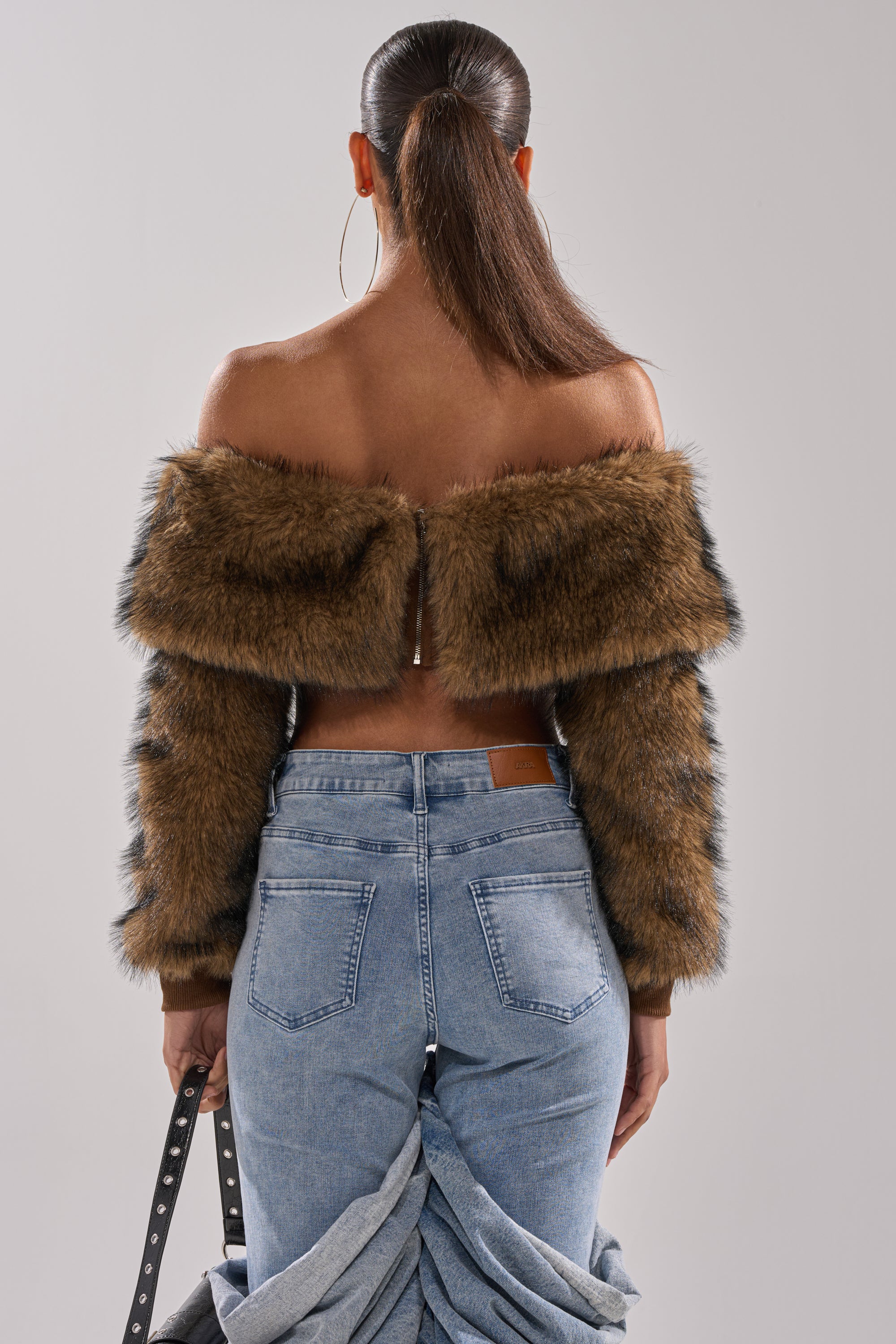 A woman faces away in the TEDDY BEAR CROPPED LONG SLEEVE TOP, paired with light blue ruffled high-waisted jeans and large hoop earrings. Holding a black strap, she embodies chic winter fashion.