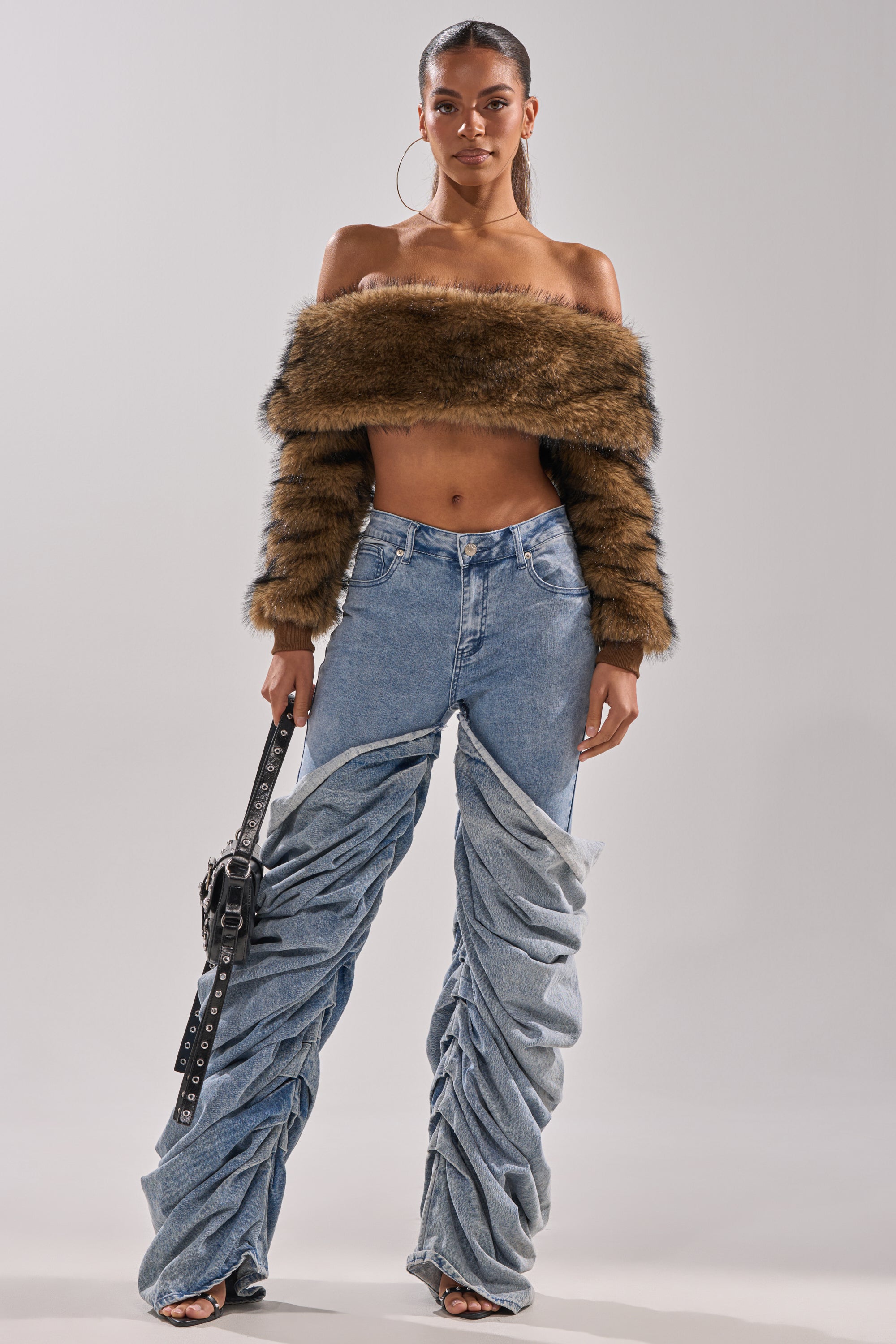 A woman faces forward wearing the TEDDY BEAR CROPPED LONG SLEEVE TOP, ruched blue denim jeans, hoop earrings, and holding a black belt-like accessory against a plain, light background.