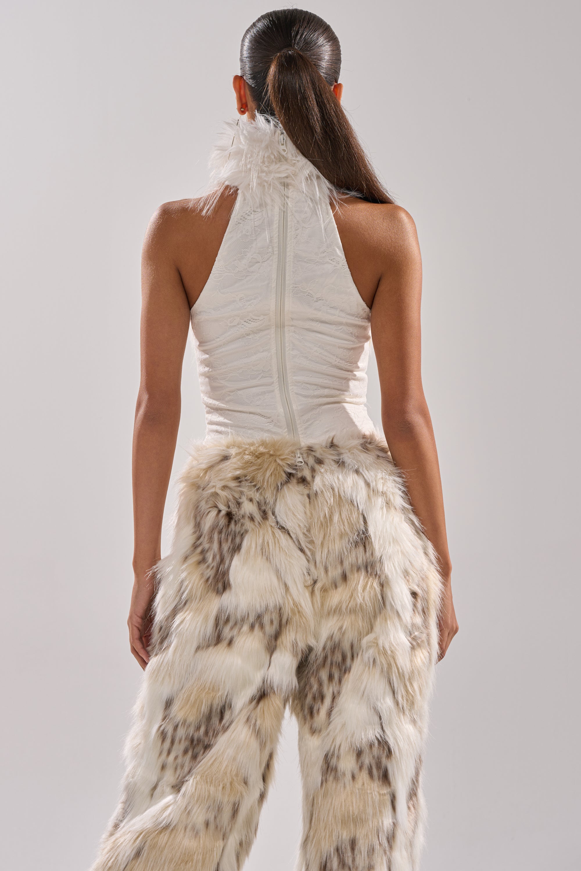 A person with dark hair in a low ponytail faces away, wearing the FLINTSTONES MOCK NECK FUR TOP and fluffy, light brown and white faux fur pants against a plain, light background.