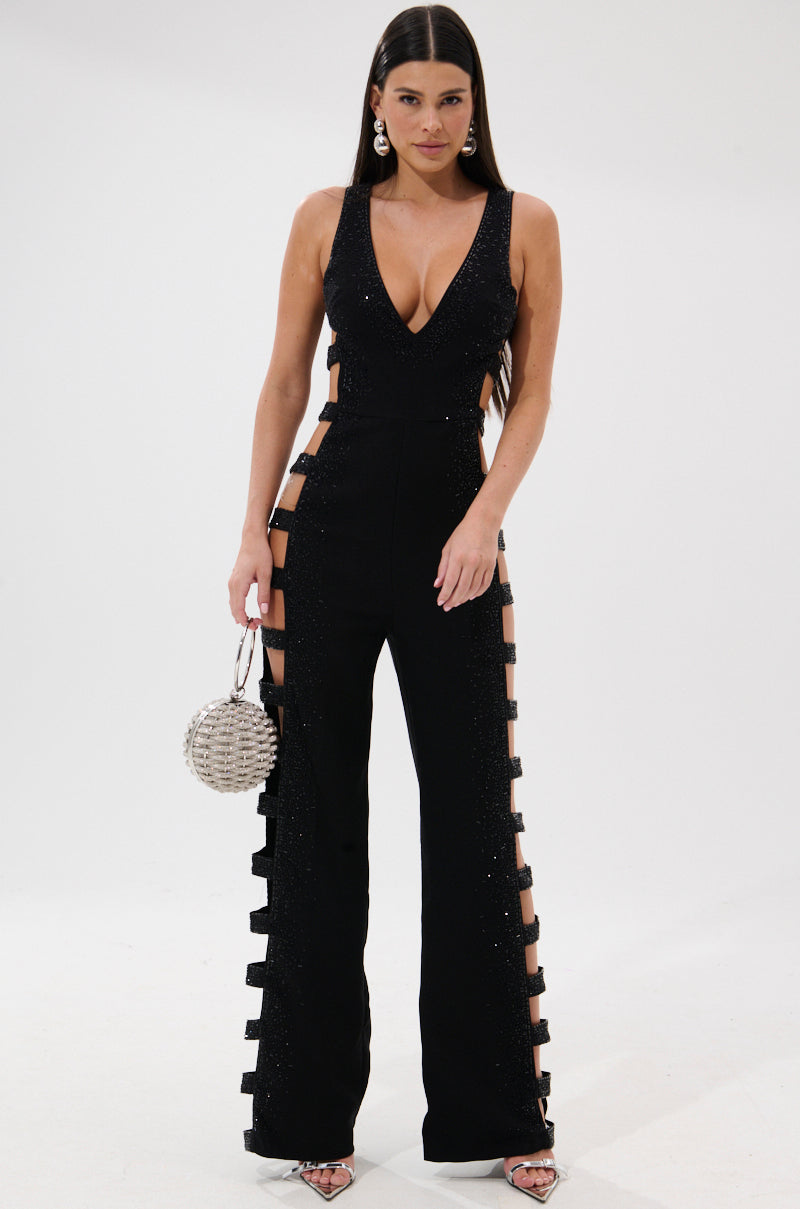 A woman wears the YOUR MAIN ONE CUTOUT JUMPSUIT WITH RHINESTONES, featuring deep side cutouts and bows, paired with a silver round handbag, large earrings, and high heels against a white background.