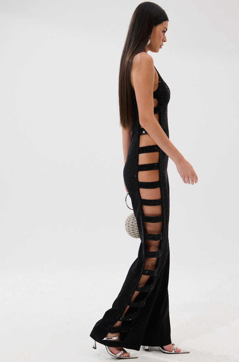 A woman with long straight hair models the YOUR MAIN ONE CUTOUT JUMPSUIT WITH RHINESTONES, paired with black heels and a round silver handbag, walking against a plain white background.