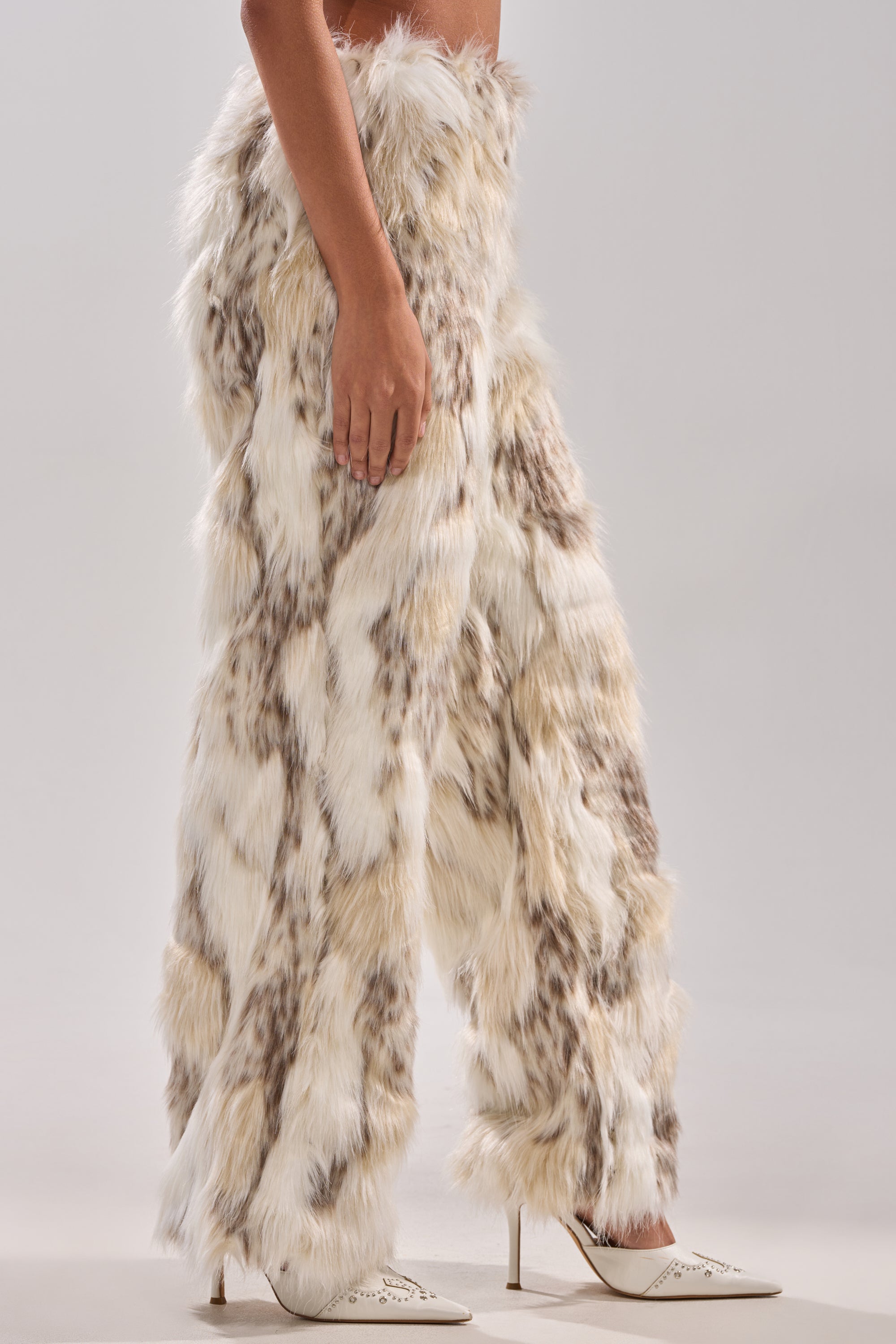 Someone stands against a plain light background, wearing FLINTSTONES FUR PANTS—high-waisted, wide-legged pants made of white and brown faux fur—paired with white pointed-toe high heels.