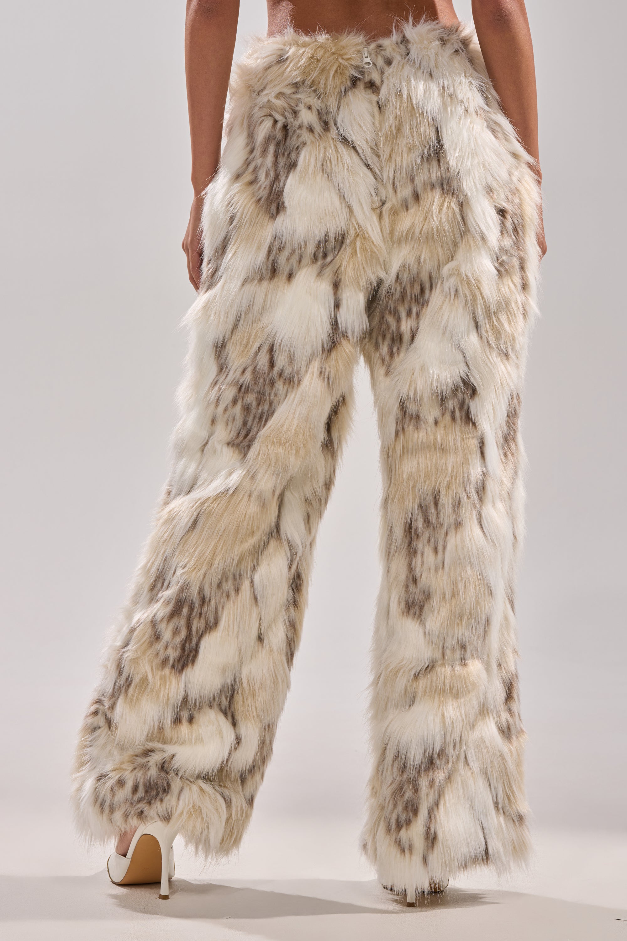 A person stands with their back to the camera, wearing FLINTSTONES FUR PANTS—wide-legged, high-waisted faux fur pants in patchy cream, beige, and brown—paired with white heels against a plain background.