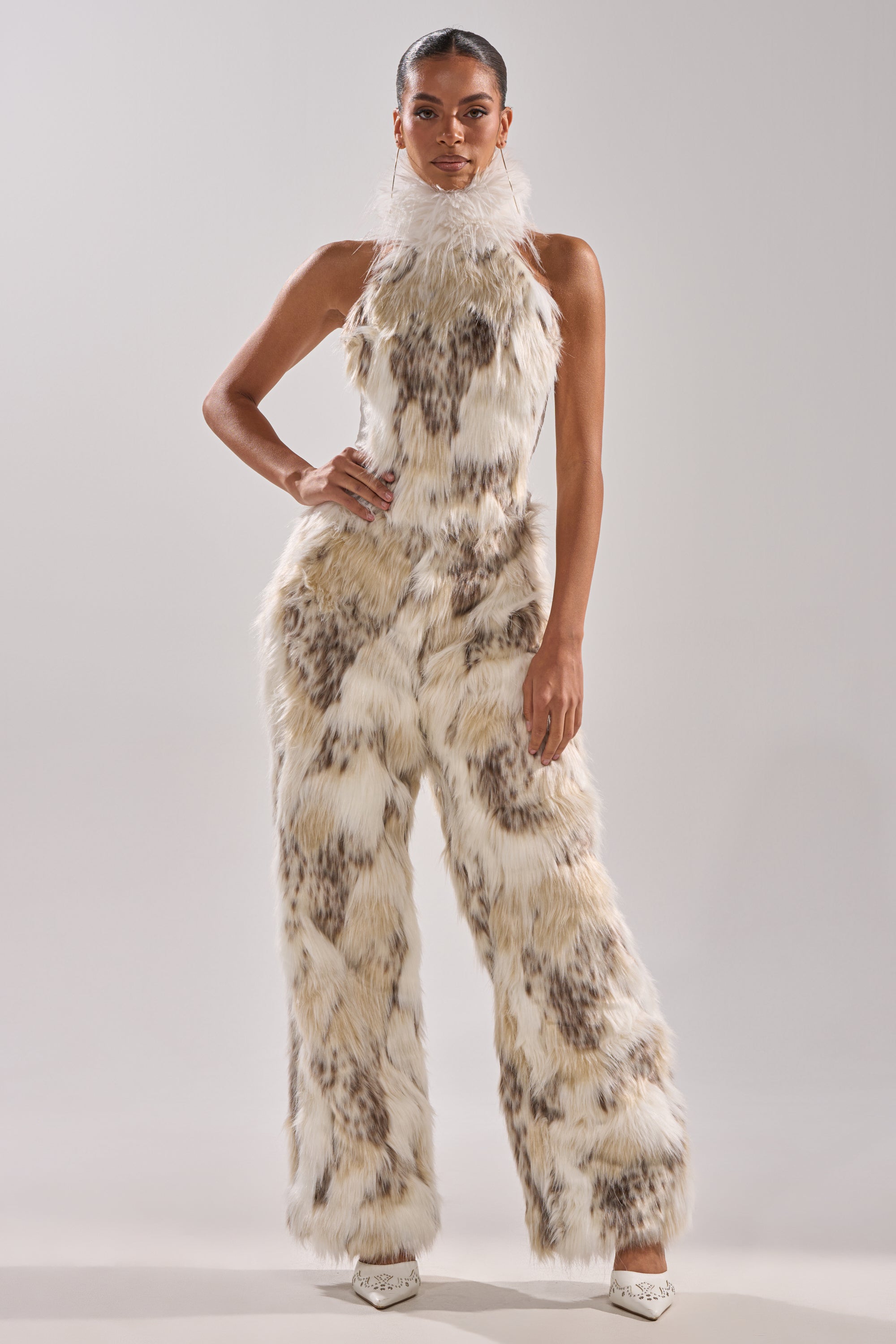 A woman poses confidently in high-waisted, wide-leg FLINTSTONES FUR PANTS, featuring white and brown faux fur for a retro look as she stands with one hand on her hip against a plain background.