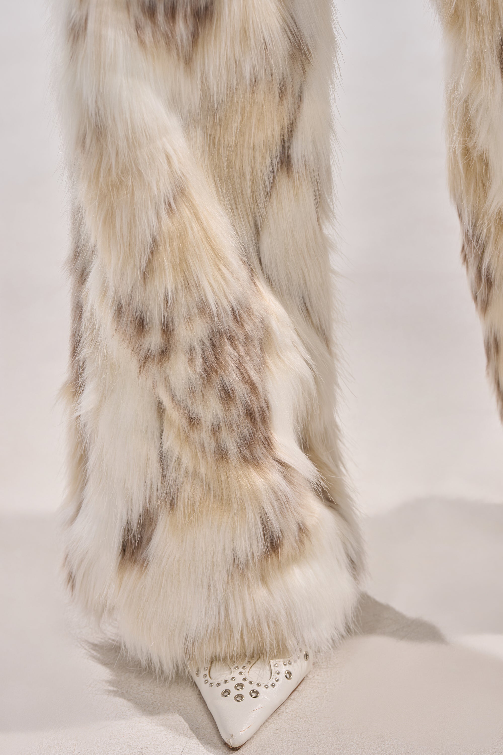 A close-up of a person wearing FLINTSTONES FUR PANTS—high-waisted faux fur in cream with brown spots—partially covering a pointed white shoe with silver studs, against a soft, light-colored background.