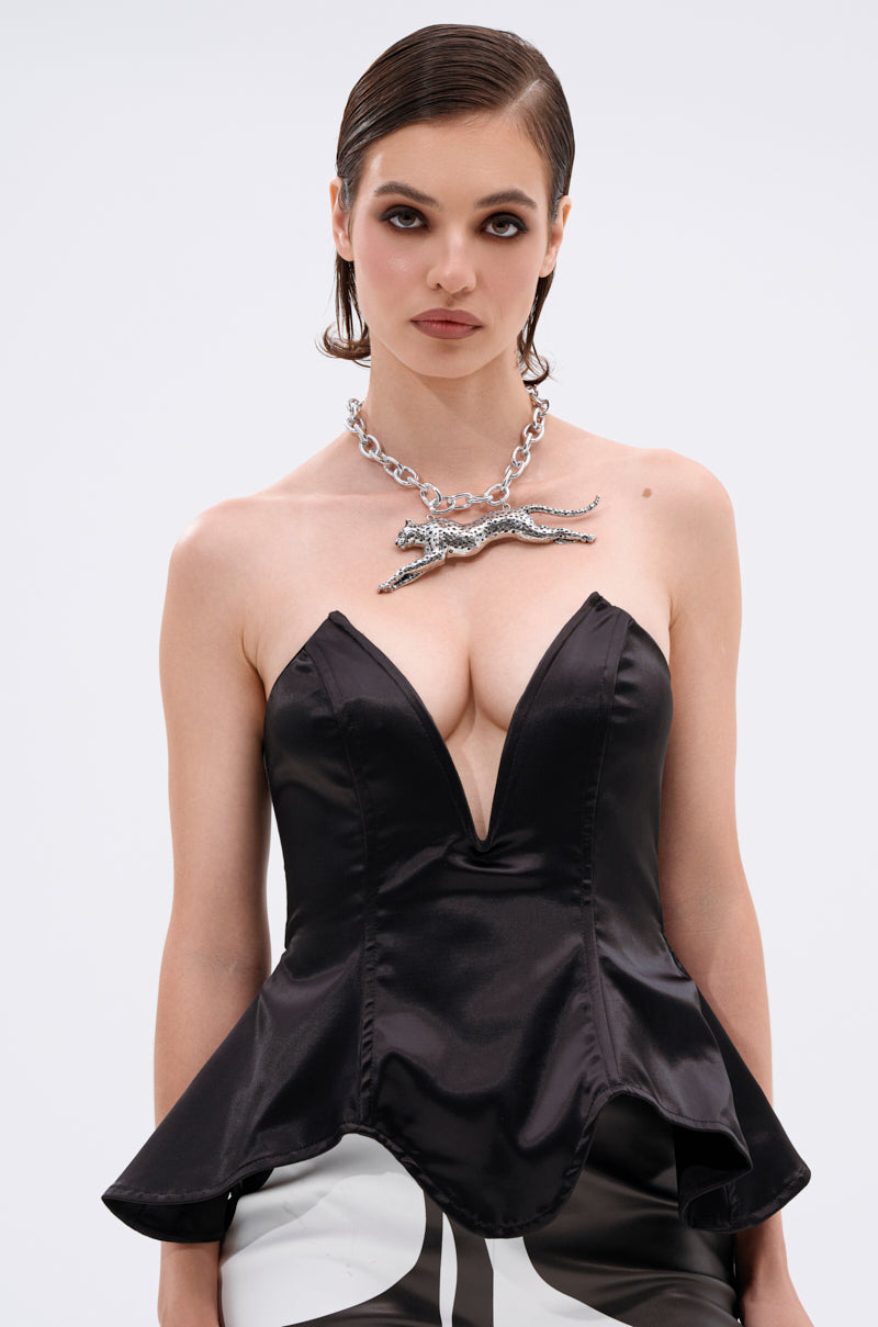 A woman with slicked-back hair wears the DRAMA QUEEN CORSET, a black satin, strapless top with a deep V neckline and flared peplum silhouette, accessorized with a bold silver chain necklace featuring an animal pendant against a white background.