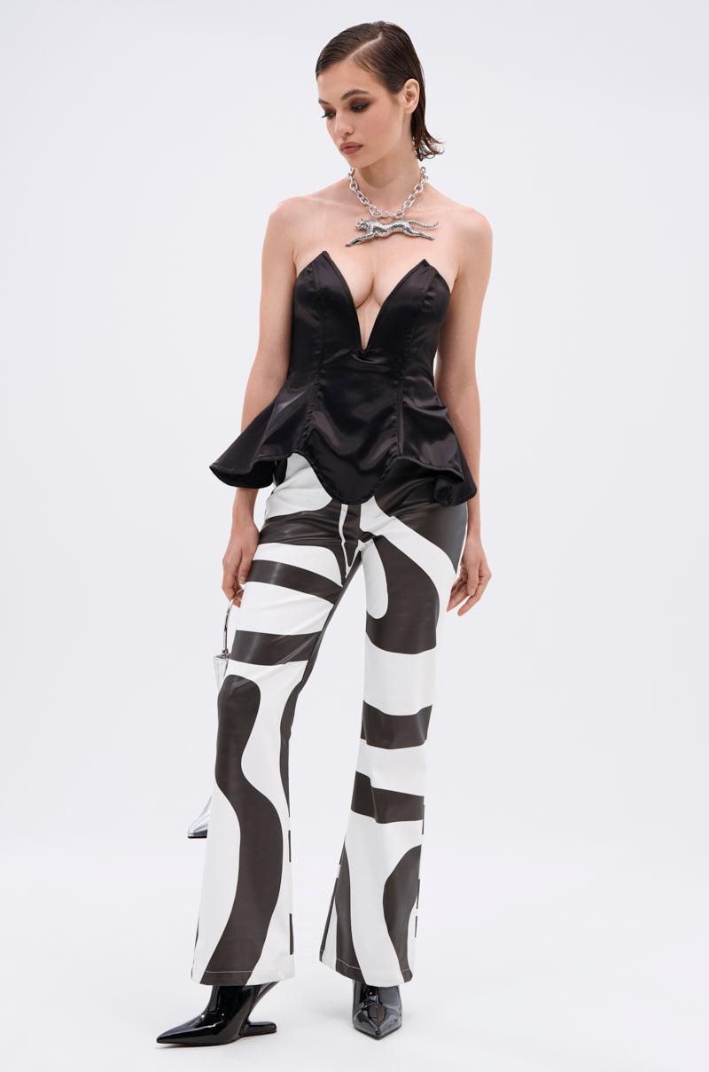 A woman stands against a plain background wearing the DRAMA QUEEN CORSET, a strapless black peplum silhouette top with a deep V neckline, paired with black and white abstract patterned pants, pointed-toe boots, and a chunky silver necklace.