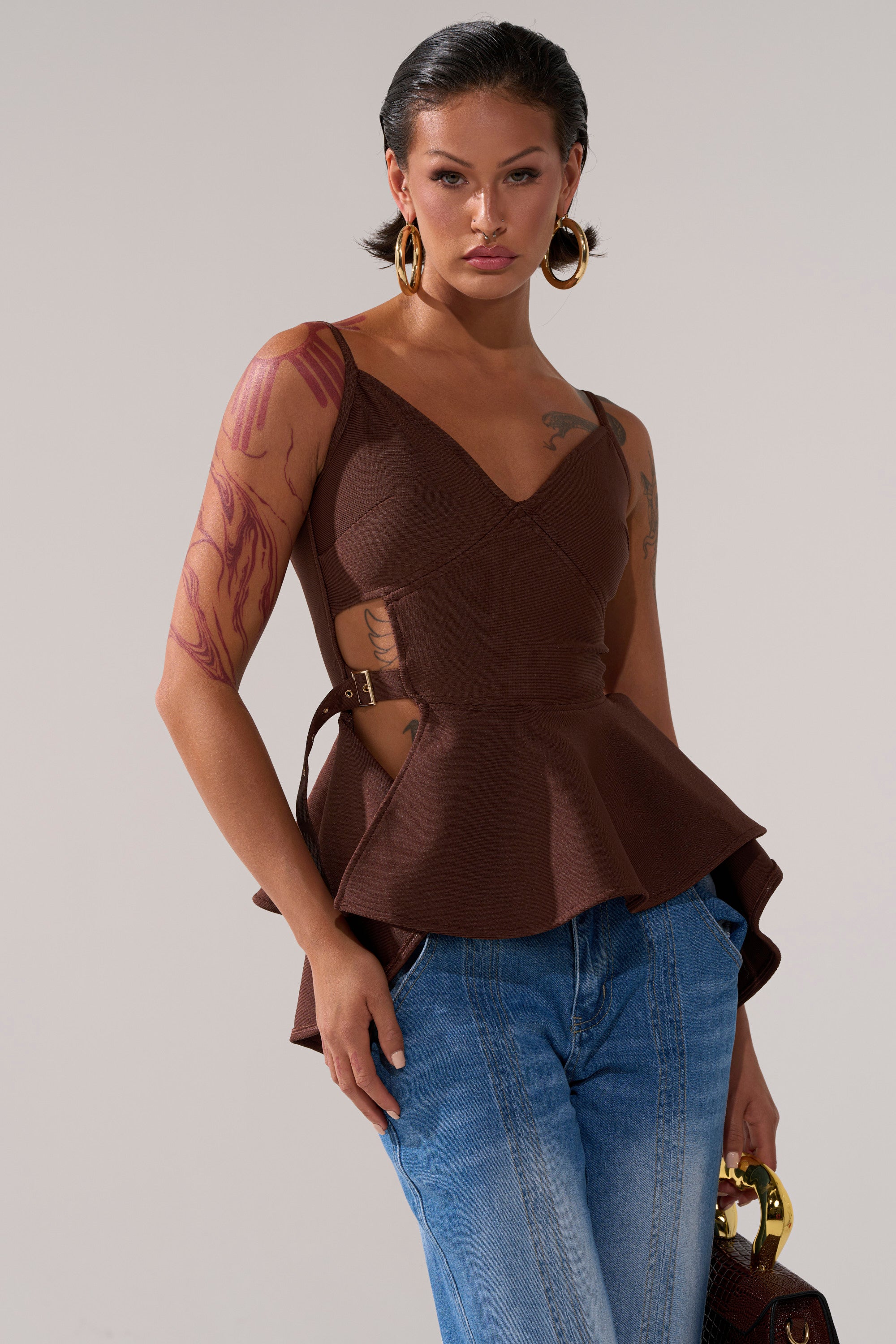 A woman with wet-look dark hair wears the TRACY SIDE ADJUSTER PEPLUM TOP in brown, styled with blue jeans, large gold hoop earrings, and a small handbag. She poses confidently, showing tattoos on her arm against a neutral background.