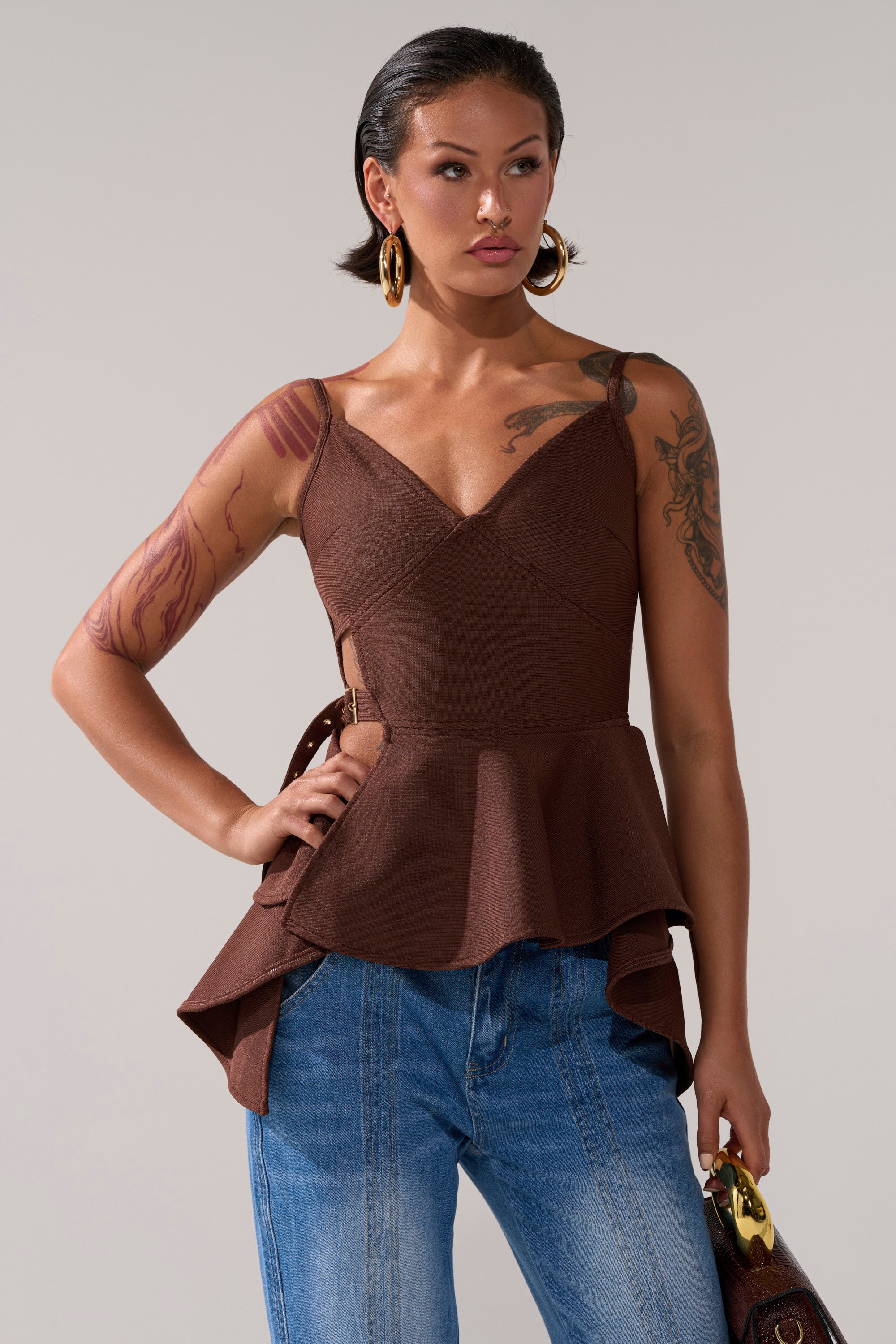 A woman with tattoos and gold hoop earrings wears the TRACY SIDE ADJUSTER PEPLUM TOP in brown, styled with blue jeans and a matching handbag. She stands against a plain background, one hand on her hip.