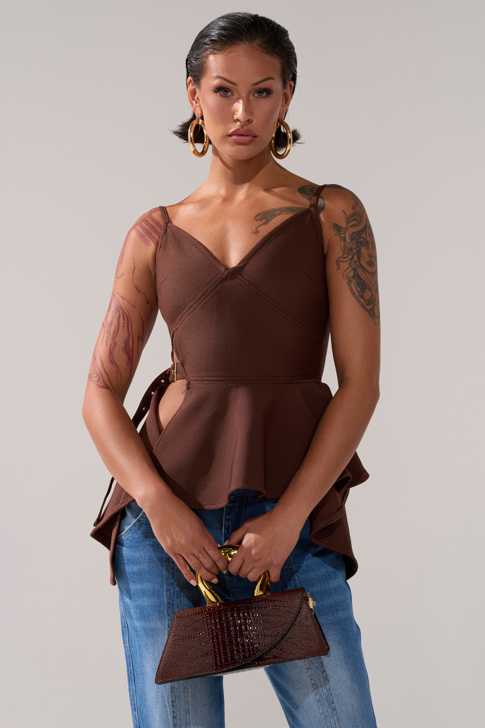 A tattooed woman with slicked-back hair and large hoop earrings wears the TRACY SIDE ADJUSTER PEPLUM TOP in brown paired with blue jeans. She holds a small brown handbag with gold handles and has a neutral expression.