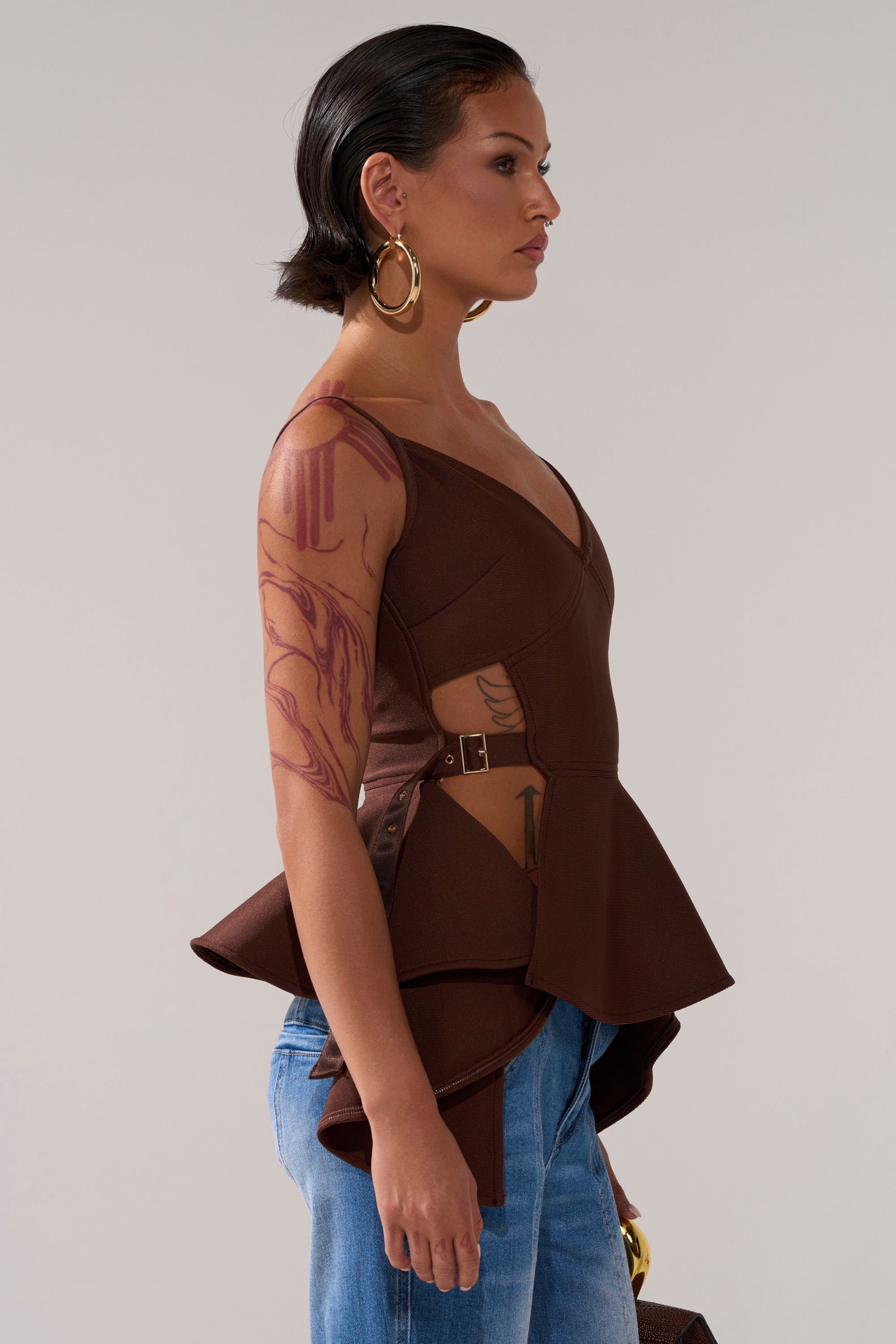A person with short dark hair, large hoop earrings, and visible arm and side tattoos wears the brown TRACY SIDE ADJUSTER PEPLUM TOP with side cutouts and blue jeans, standing in profile against a plain background.
