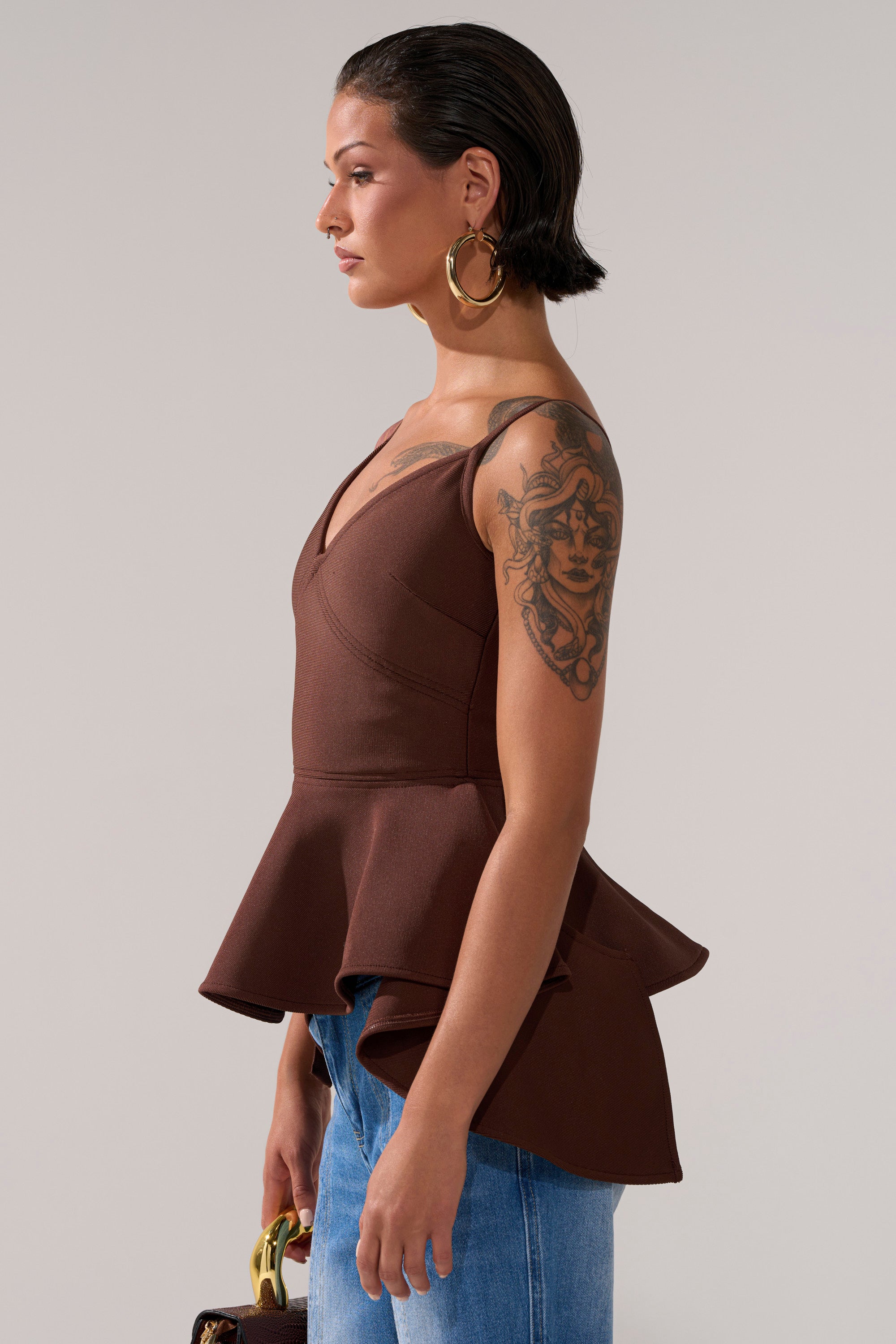 A woman with short dark hair and a prominent upper arm tattoo stands in profile wearing the TRACY SIDE ADJUSTER PEPLUM TOP in brown, paired with blue jeans and large hoop earrings, holding a small handbag.