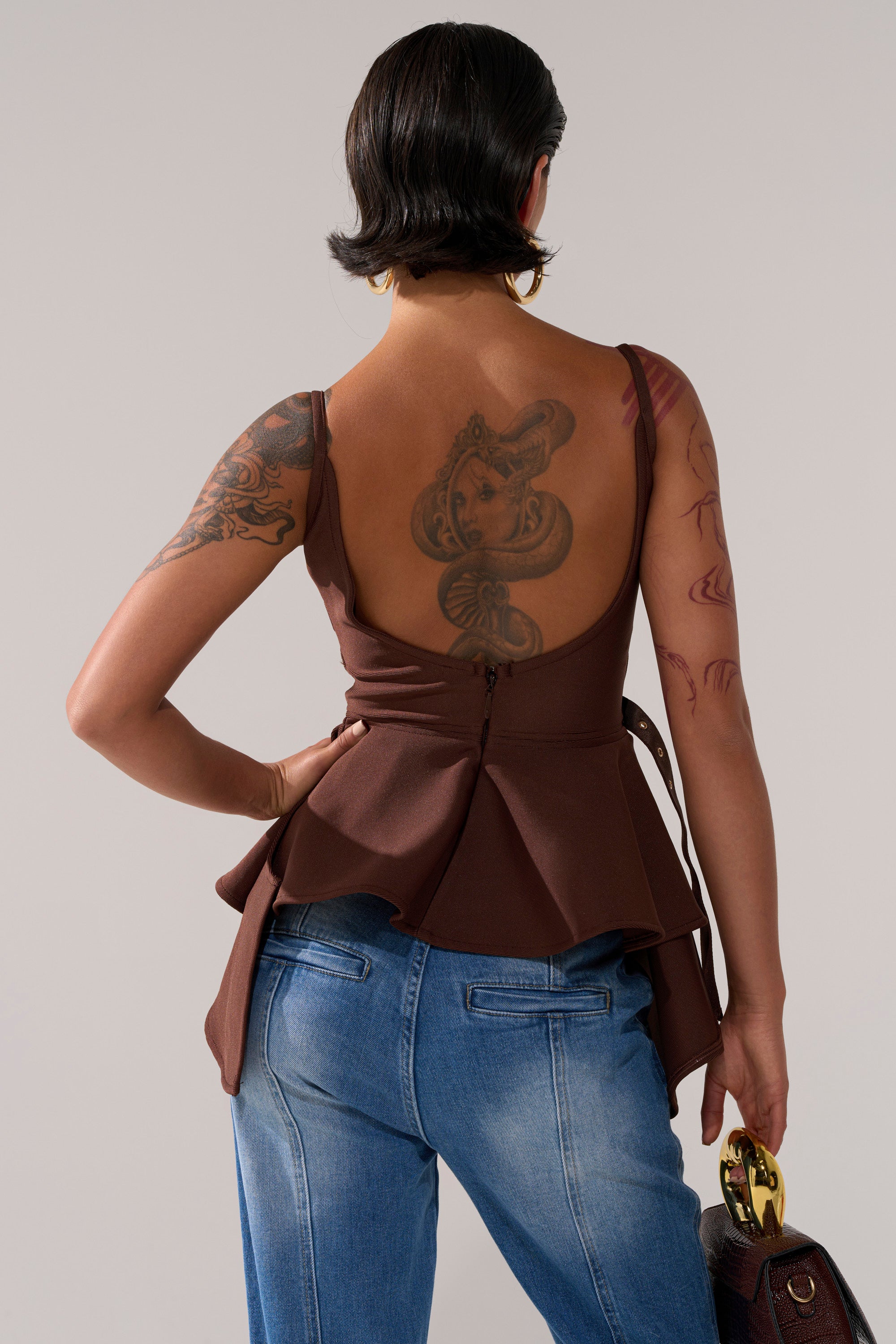 Wearing the TRACY SIDE ADJUSTER PEPLUM TOP and blue jeans, a person with short dark hair displays a large snake tattoo on their upper back and arm tattoos while carrying a brown handbag with adjustable straps.
