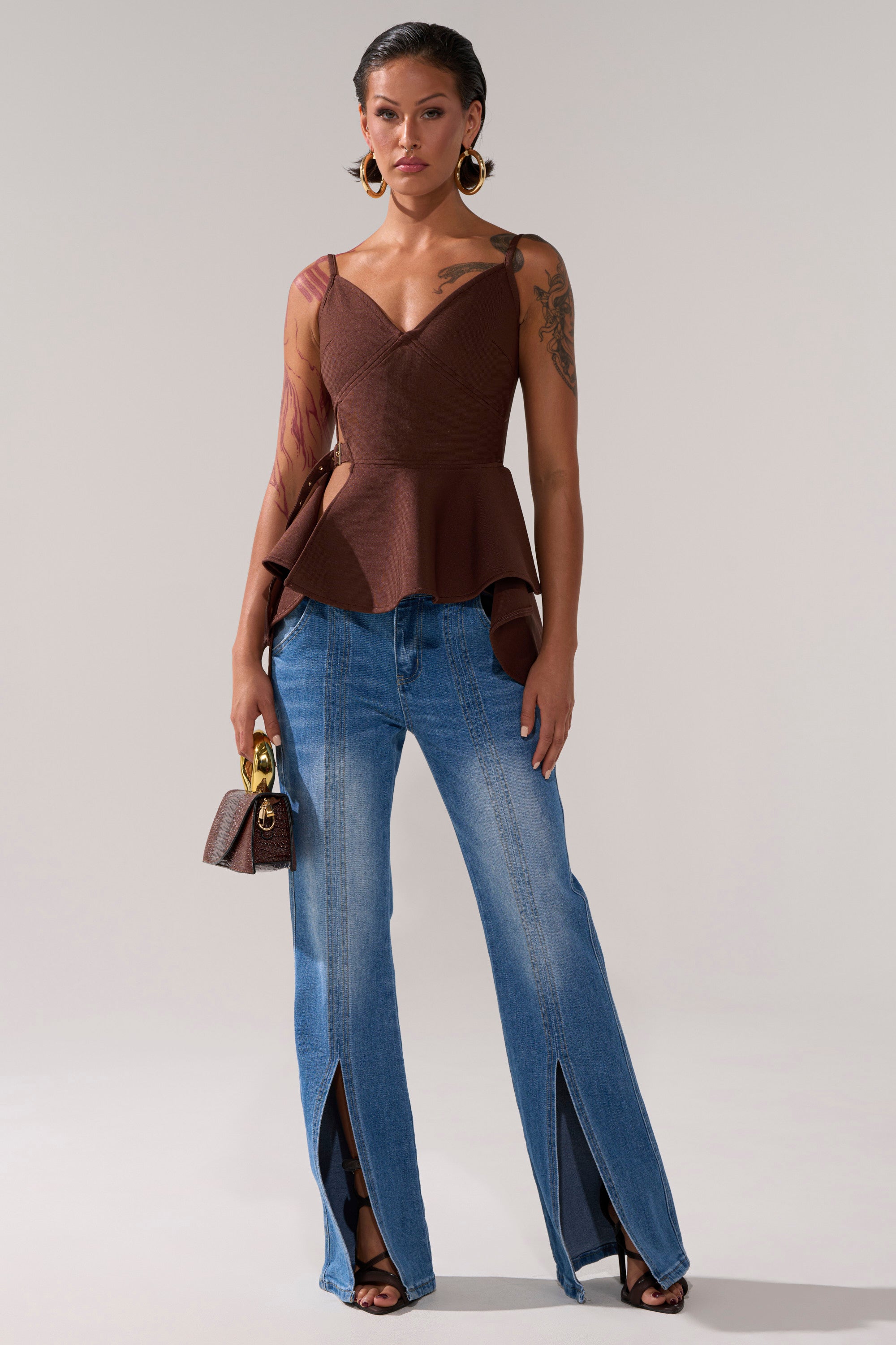 A woman in the TRACY SIDE ADJUSTER PEPLUM TOP, blue wide-leg jeans with front slits, black heeled sandals, large hoop earrings, and a small brown handbag stands against a neutral background.
