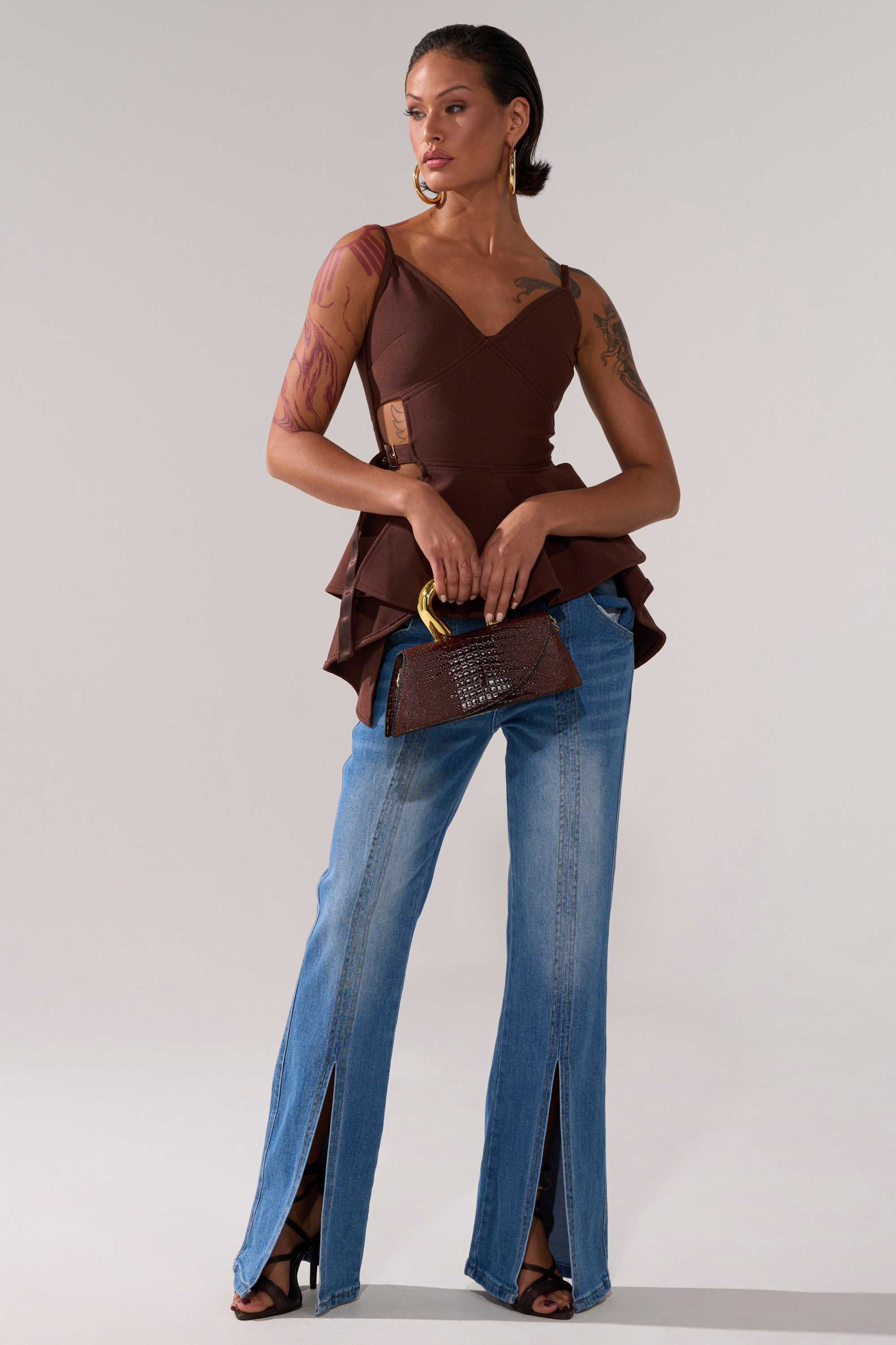 A woman wears the TRACY SIDE ADJUSTER PEPLUM TOP in brown with adjustable straps, paired with blue jeans.