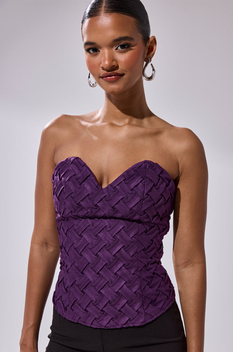 A woman with slicked-back hair models the FEELING SPECIAL SUEDE CORSET in deep purple with a sweetheart neckline and woven texture, styled with hoop earrings, posed against a neutral background.