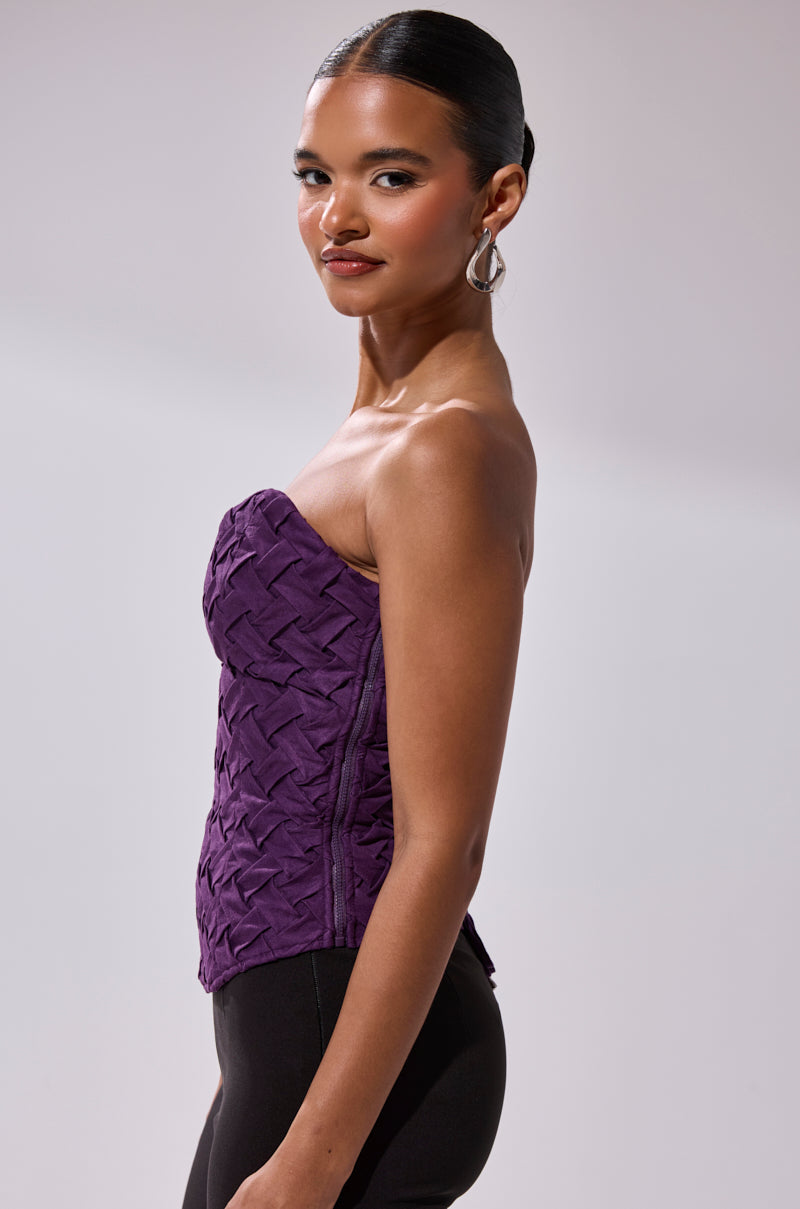 A woman with slicked-back hair wears silver hoop earrings, black pants, and the FEELING SPECIAL SUEDE CORSET—a strapless purple textured top—posing sideways and smiling slightly against a plain light background.