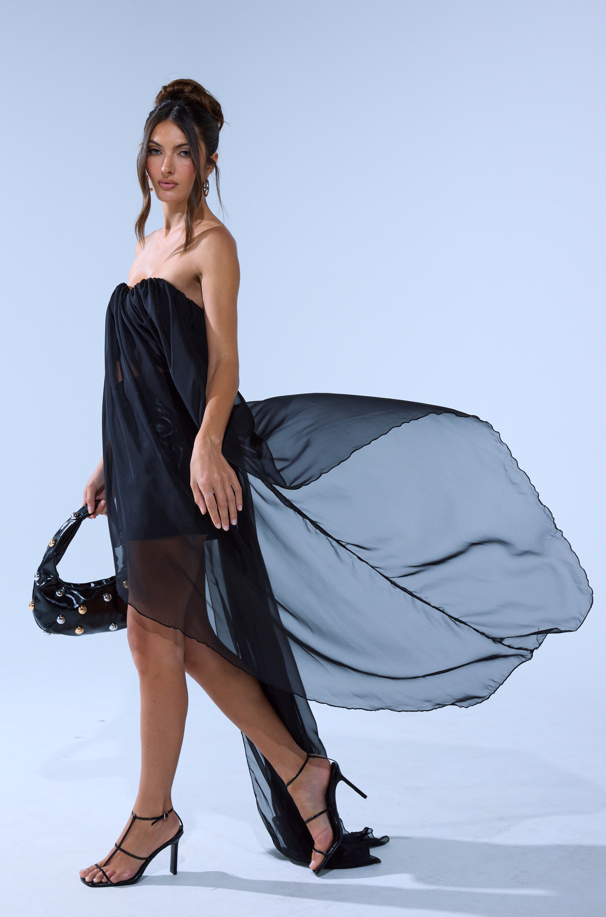 A woman in the GONE TOO LONG BLOUSE, styled with black heels and a black handbag, walks against a plain light blue background as the sheer fabric flows behind her.