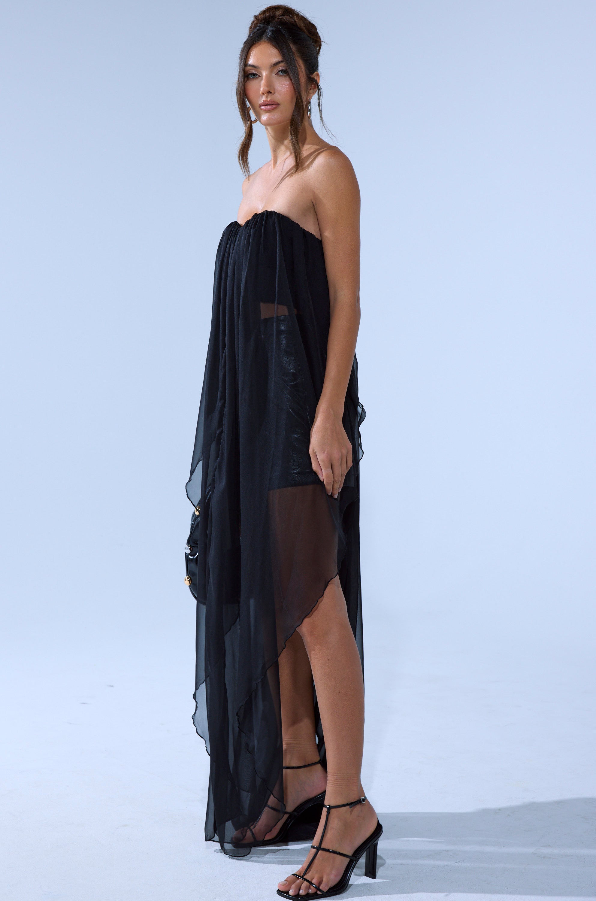 A woman stands against a light background wearing the GONE TOO LONG BLOUSE, styled with black heeled sandals and her hair up.