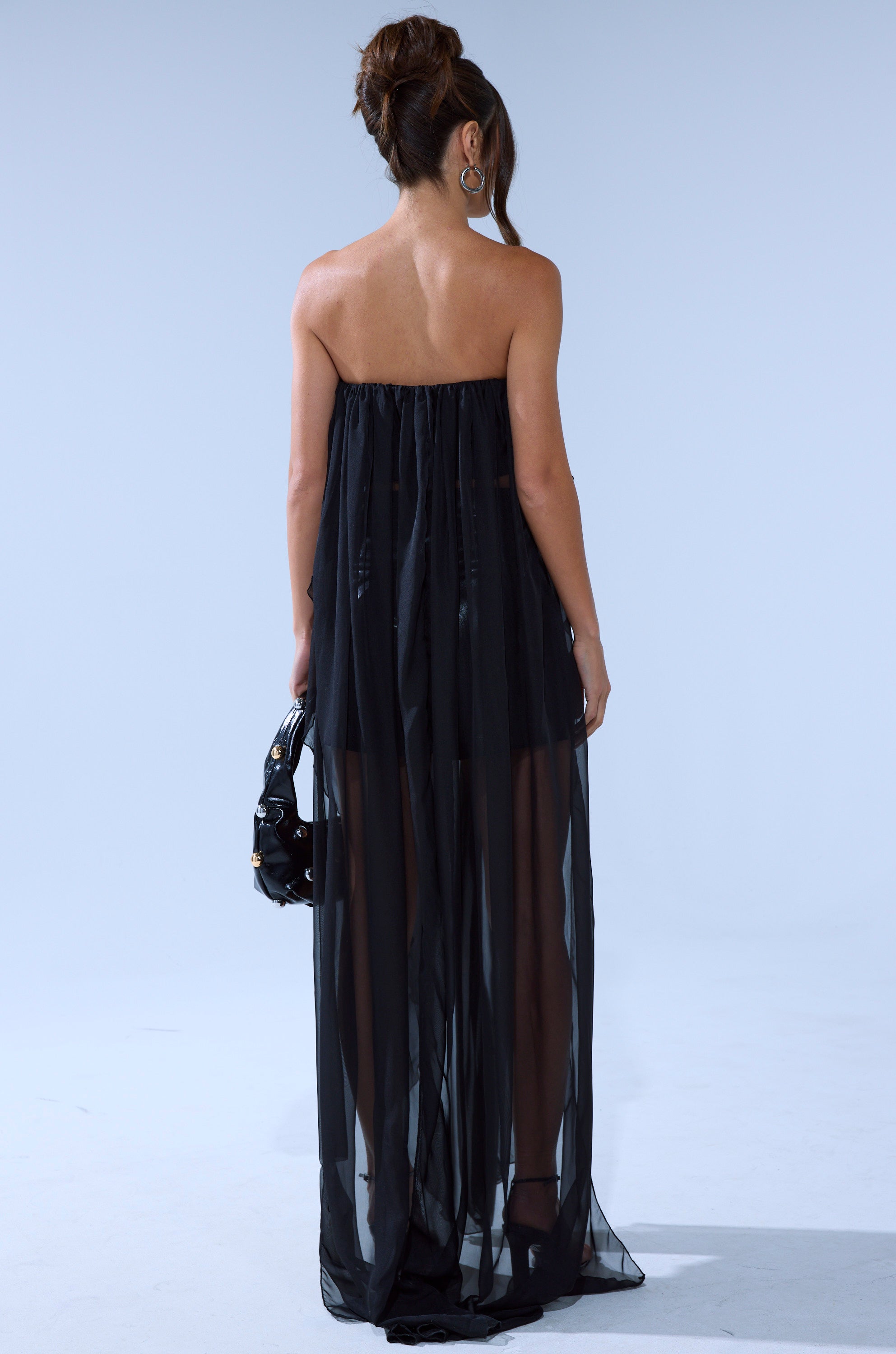 A woman stands facing away, wearing the GONE TOO LONG BLOUSE in black sheer chiffon with a short underlayer. She holds a black handbag, her hair styled in a high bun, set against a plain light background.
