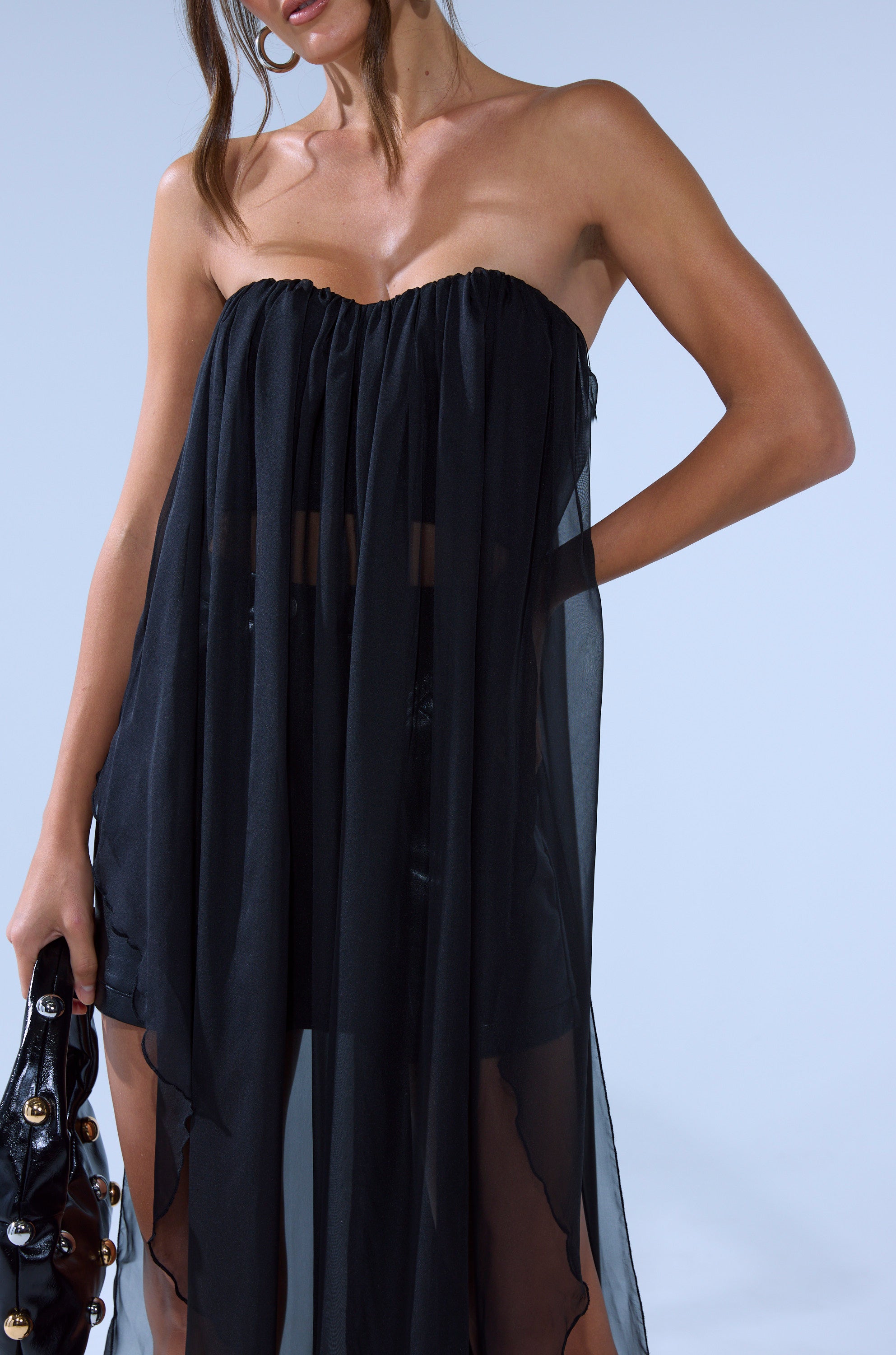 A woman, partially out of frame, with long wavy hair and hoop earrings, wears the GONE TOO LONG BLOUSE—a strapless, sheer black top with a chiffon overlay—and holds a black handbag with gold accents.