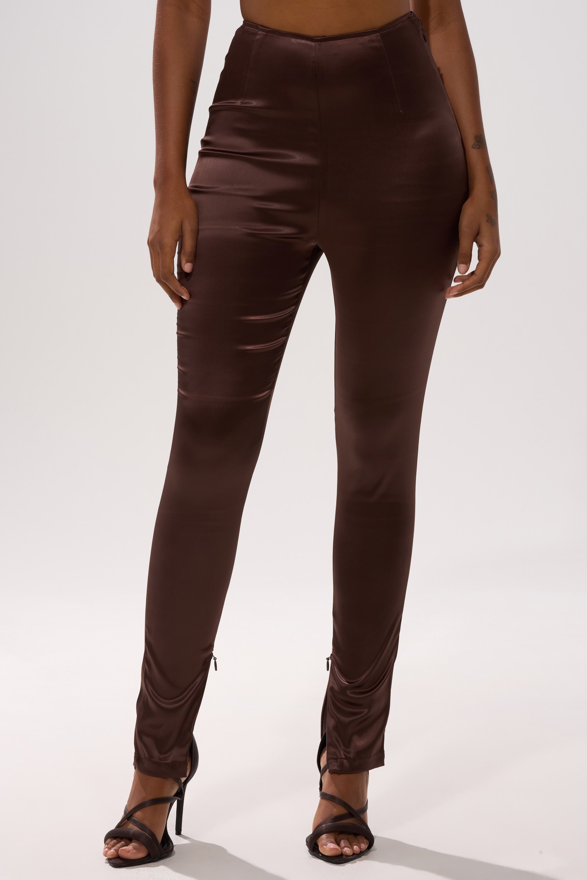 A person wearing AINT MY PROBLEM LEGGING—high-waisted, fitted, shiny brown satin leggings—and black high-heeled sandals, shown from mid-torso to feet against a plain light background.