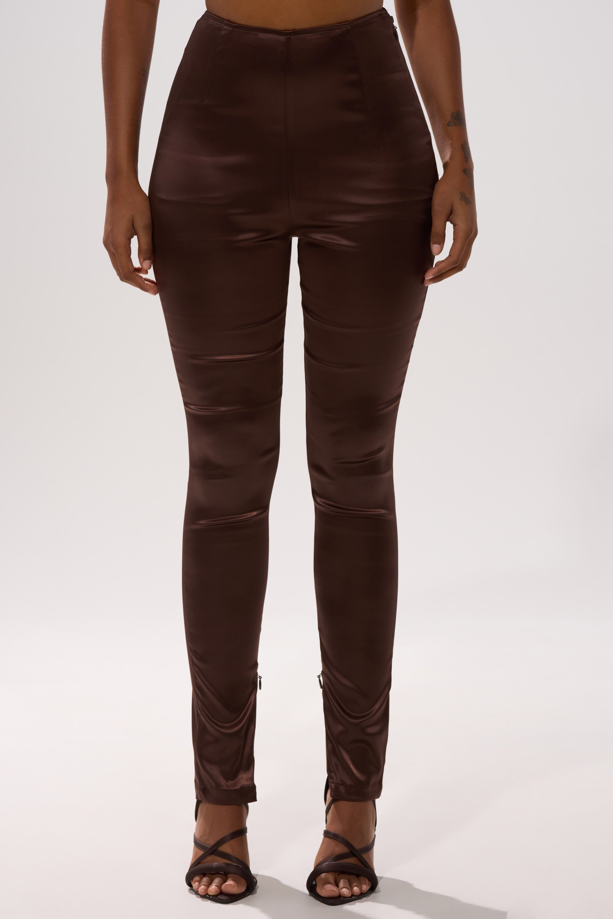 A person models the AINT MY PROBLEM LEGGING in shiny brown satin high-rise style with black strappy heels, standing against a plain light background and showing their outfit from the waist down.