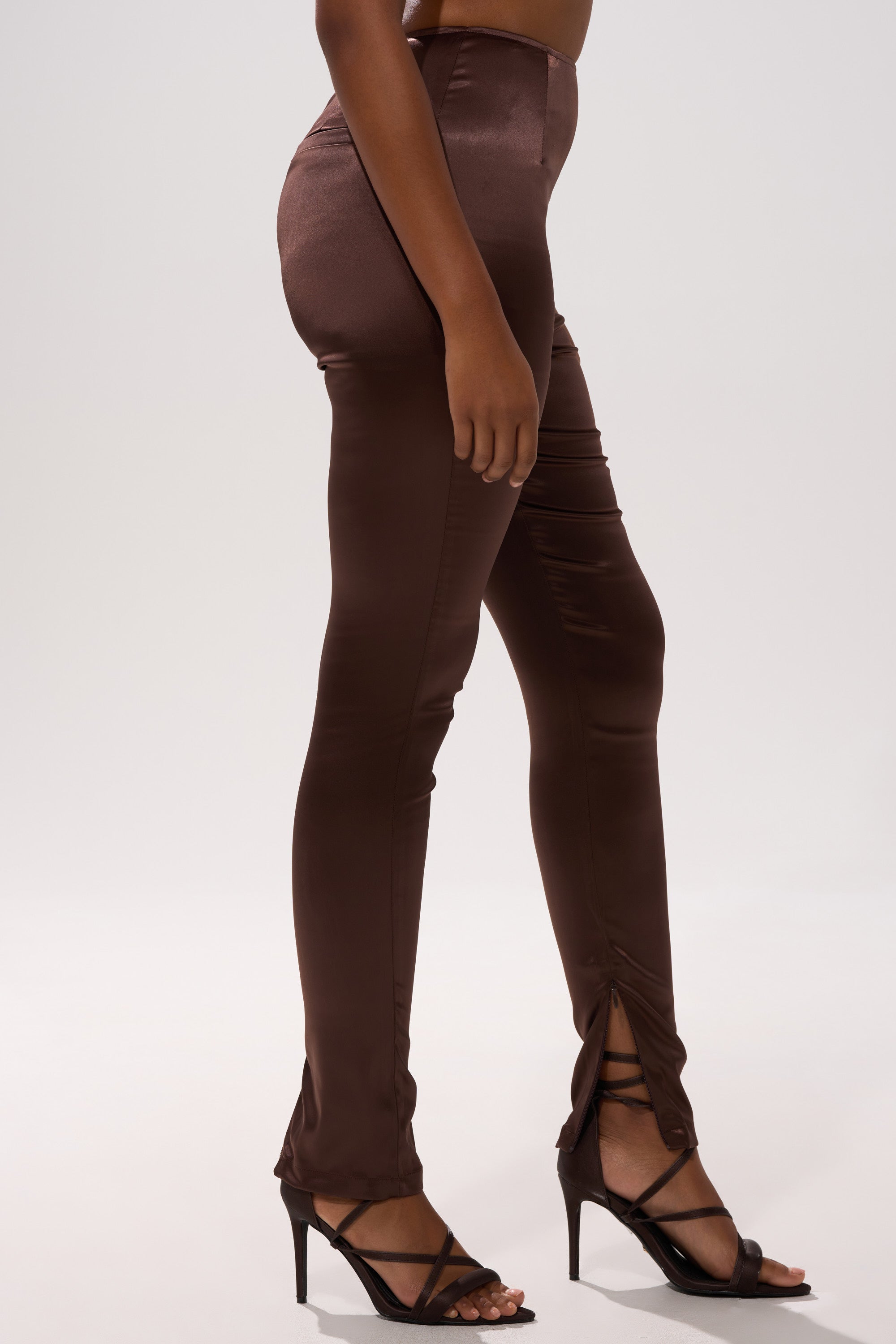 A person wearing AINT MY PROBLEM LEGGING—satin, high-rise with small ankle slits—and black strappy high-heeled sandals, standing against a plain light background.