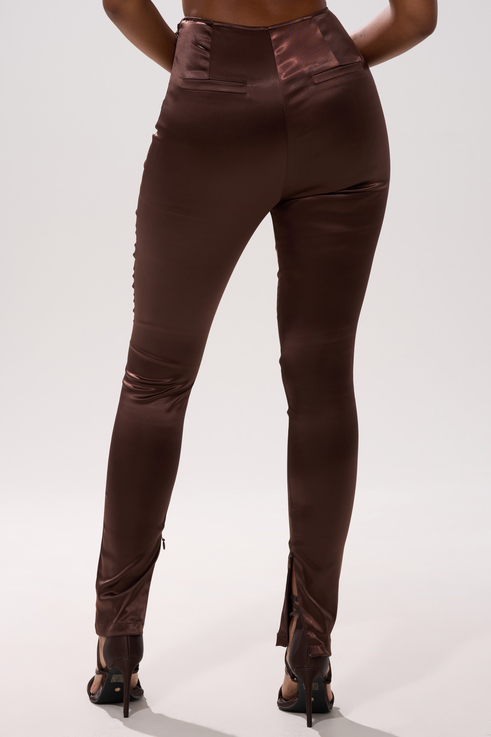 A person wearing shiny, high-waisted brown AINT MY PROBLEM LEGGING and black high-heeled sandals, shown from the back against a plain light background.