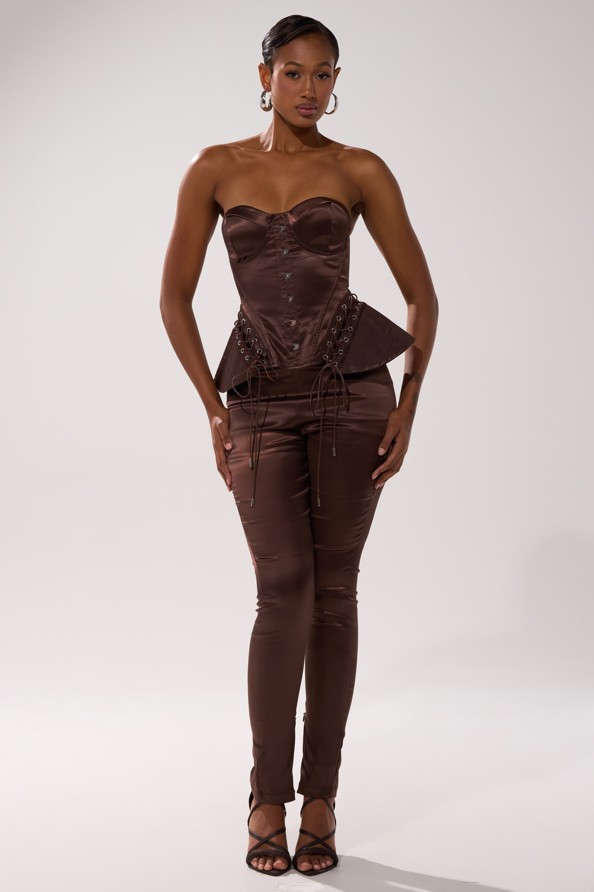 A woman stands against a plain backdrop in a strapless dark brown lace-up corset top, AKIRA Label AINT MY PROBLEM LEGGING, hoop earrings, slicked-back hair, and black strappy heels.