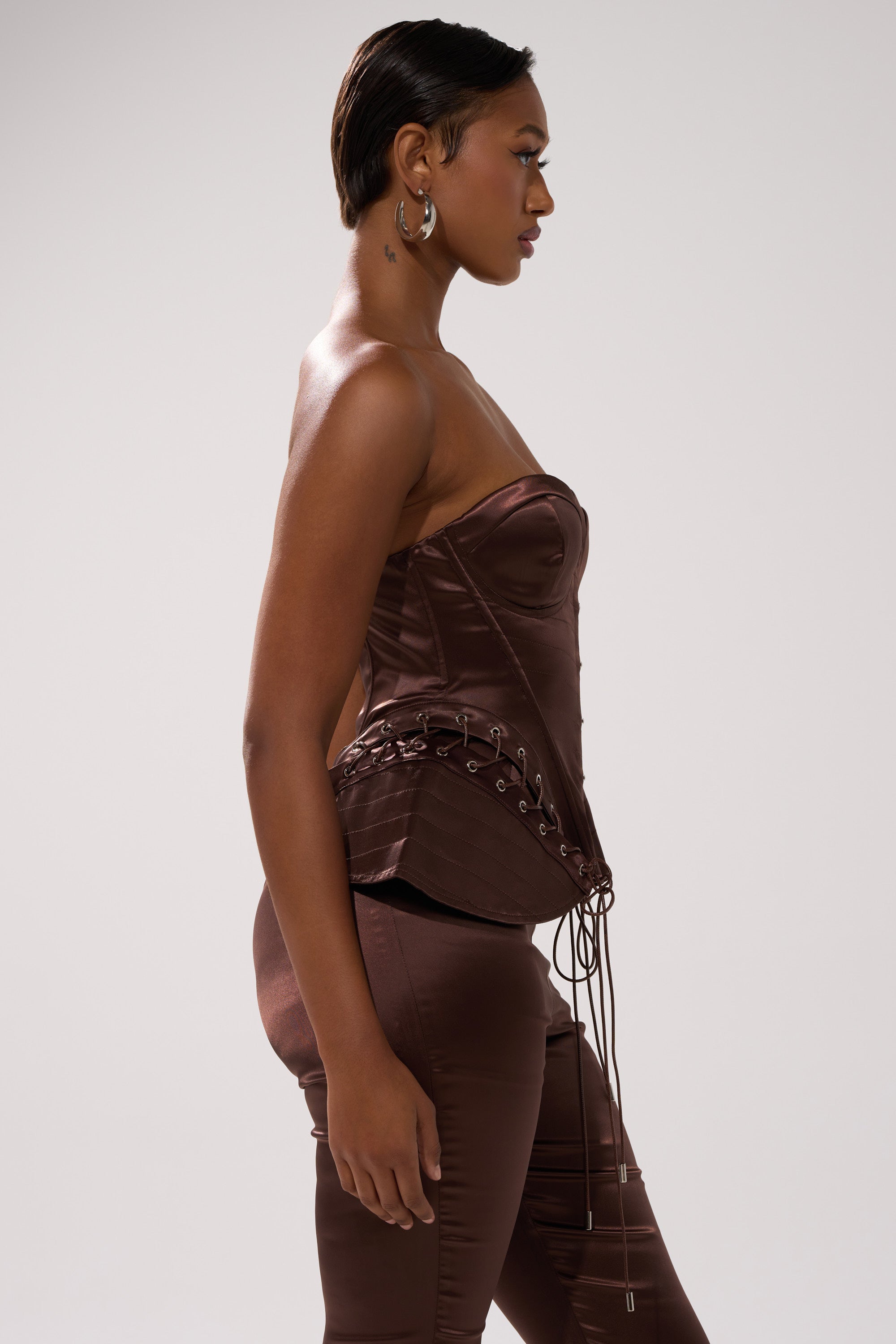 A woman in the AINT MY PROBLEM CORSET—a fitted, strapless satin top with lace-up detailing—paired with matching pants poses in profile against a plain light background. She has short, slicked-back hair and hoop earrings.
