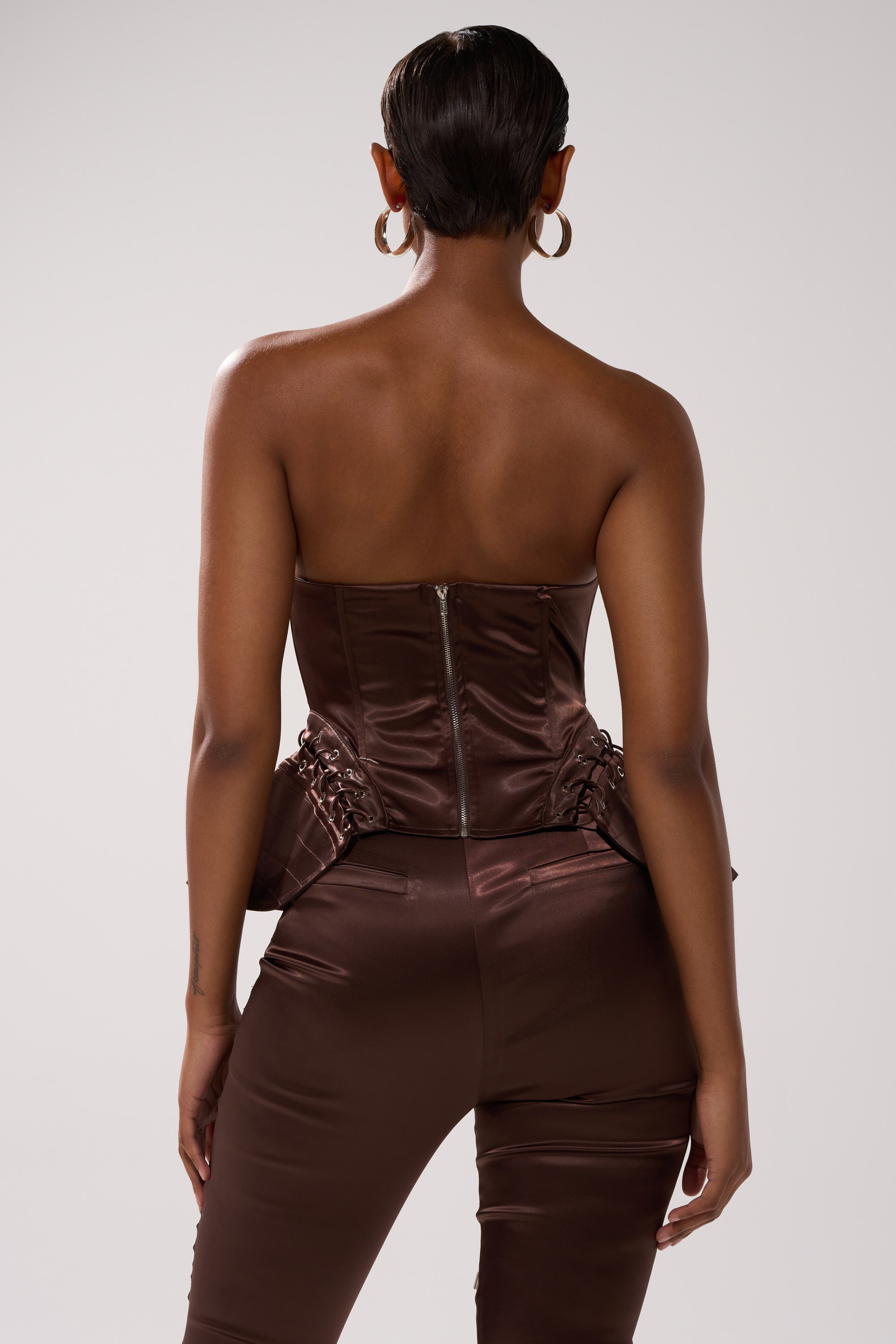 A person with short hair and hoop earrings is seen from the back wearing the AINT MY PROBLEM CORSET, a strapless, fitted satin top with a visible back zipper, subtle lace-up detailing, and matching high-waisted pants.