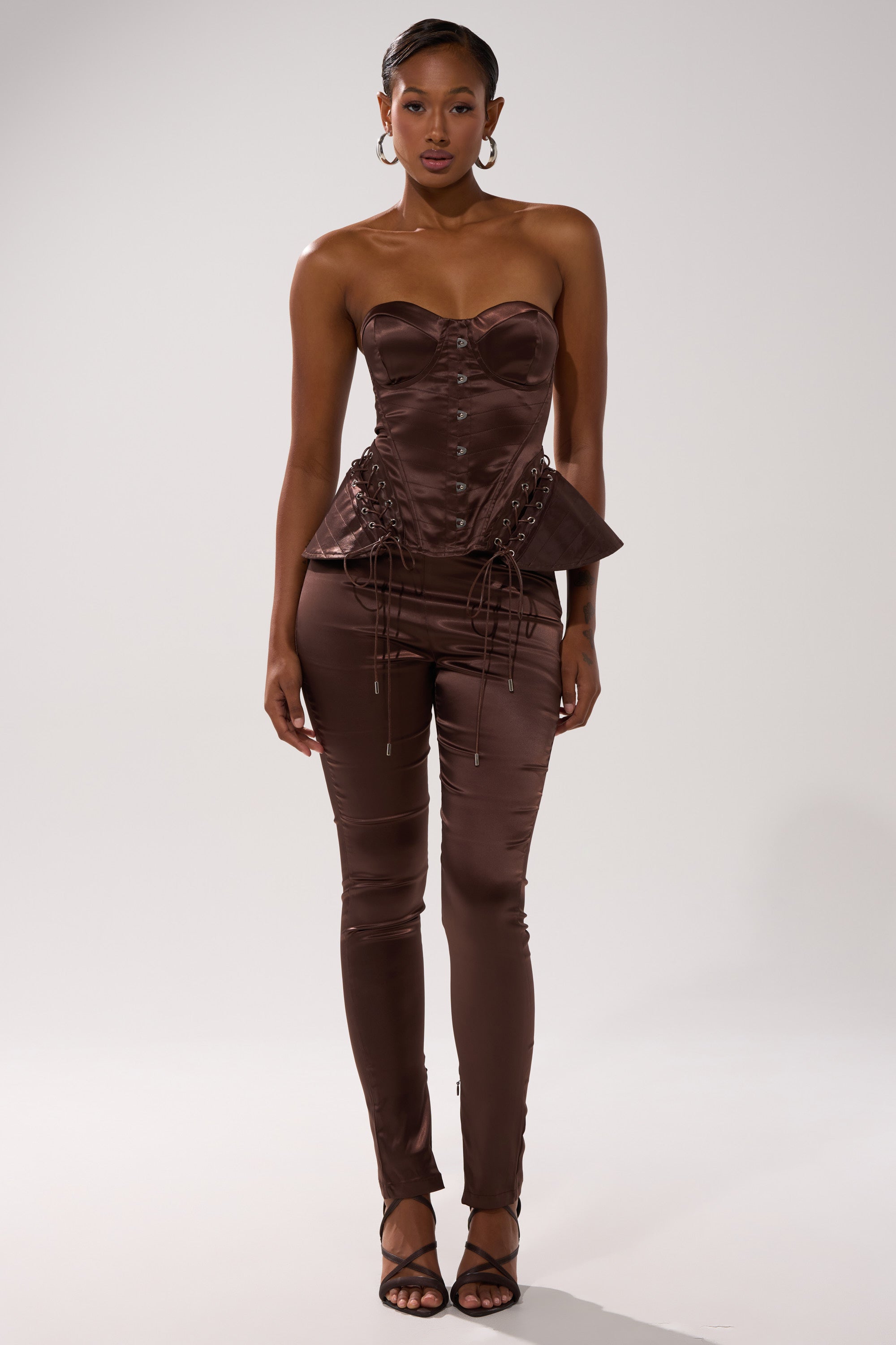 A woman faces forward in the AINT MY PROBLEM CORSET, a shiny chocolate brown satin top with lace-up detailing, paired with matching fitted pants. She wears hoop earrings, strappy heels, and her hair is pulled back against a plain light background.