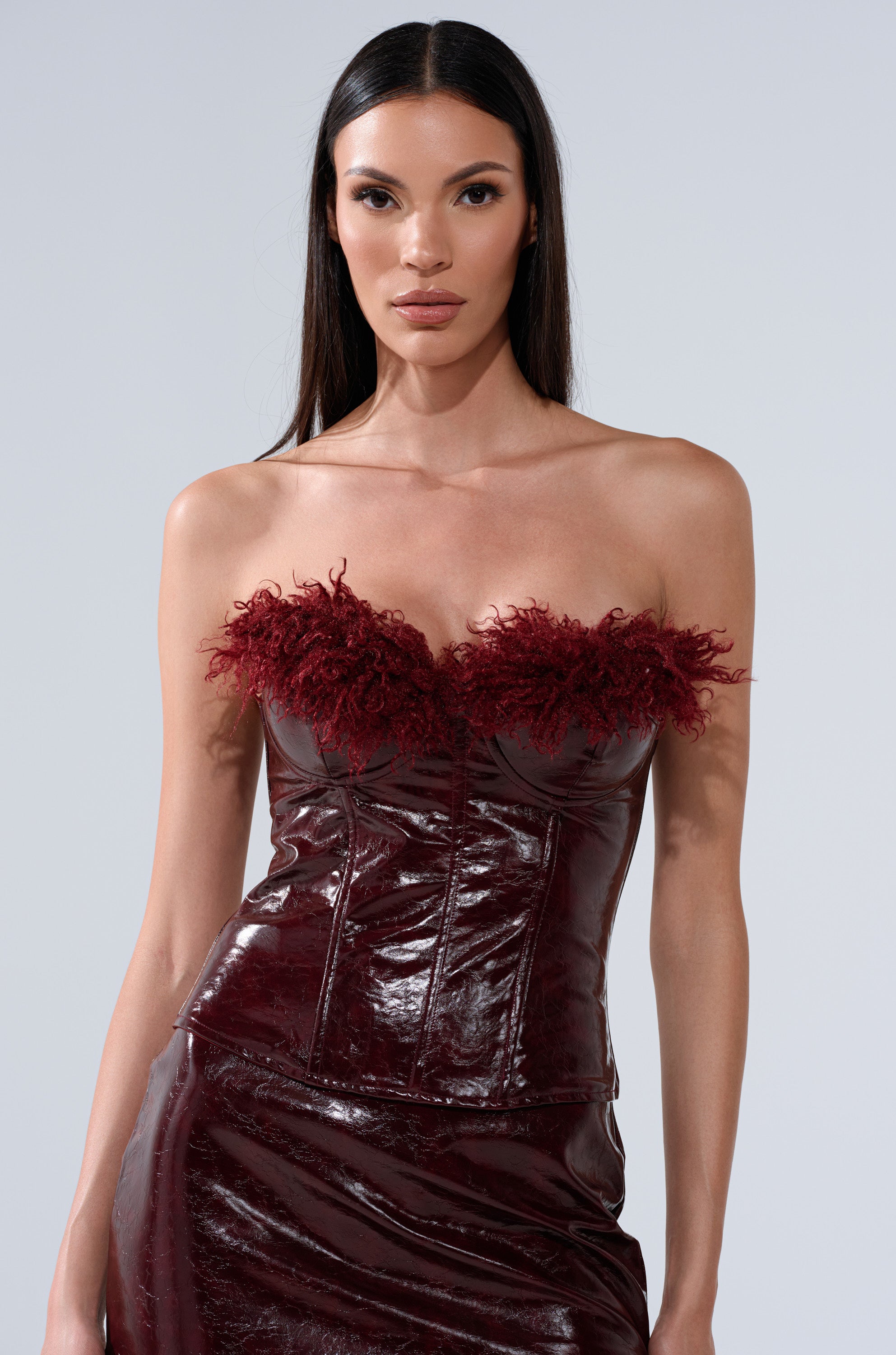 A woman with long dark hair models the "IM YOUR WEAKNESS FAUX LEATHER CORSET WITH FUR," a strapless burgundy dress with a faux leather corset bodice and feathered neckline, against a plain light gray background.